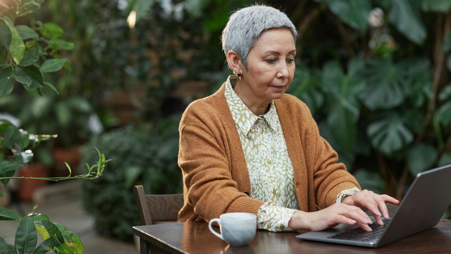 An elderly woman using a laptop outdoors surrounded by lush green plants.