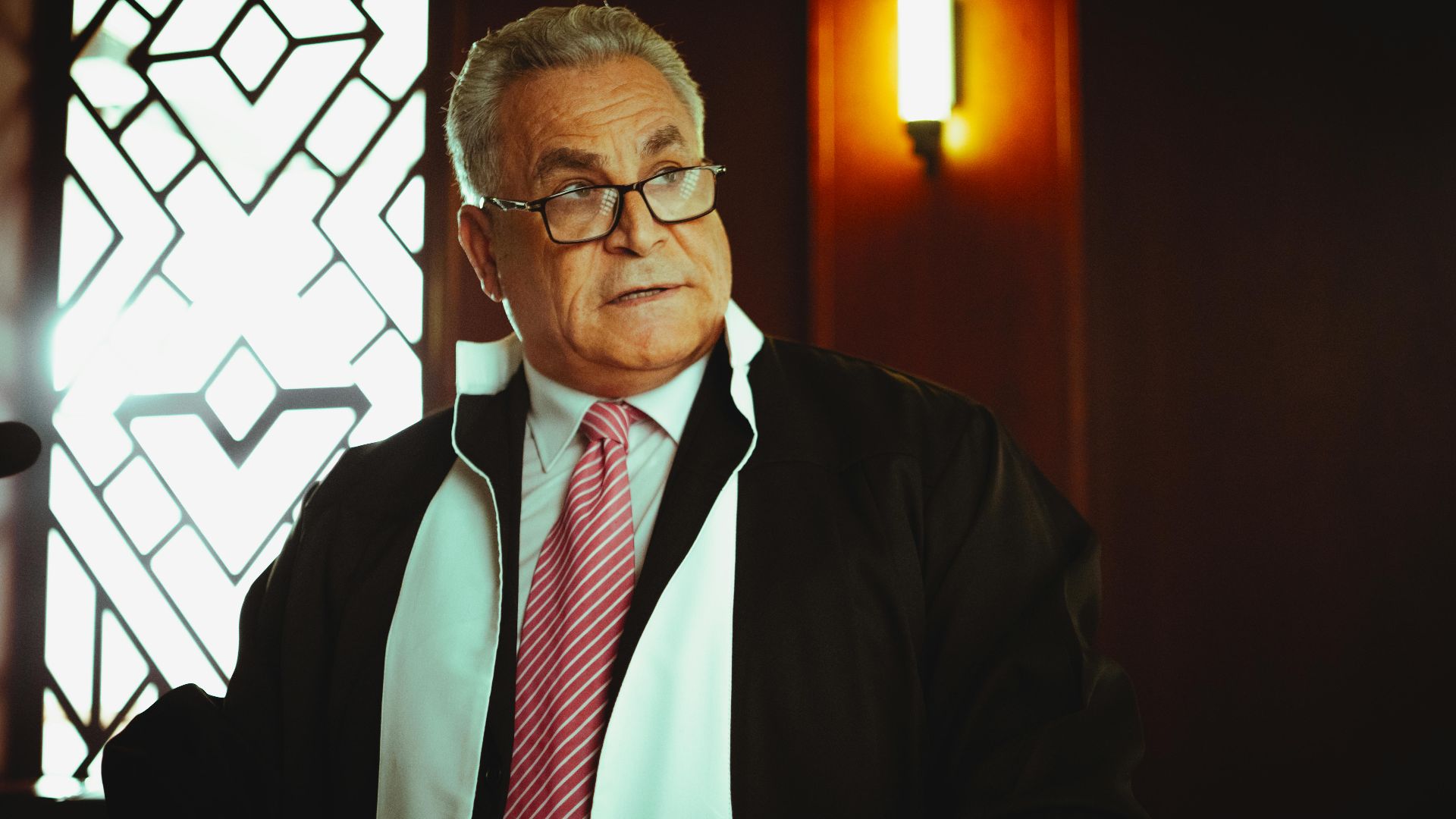 A male judge in a courtroom in Baghdad wearing a robe and tie, in a contemplative pose.