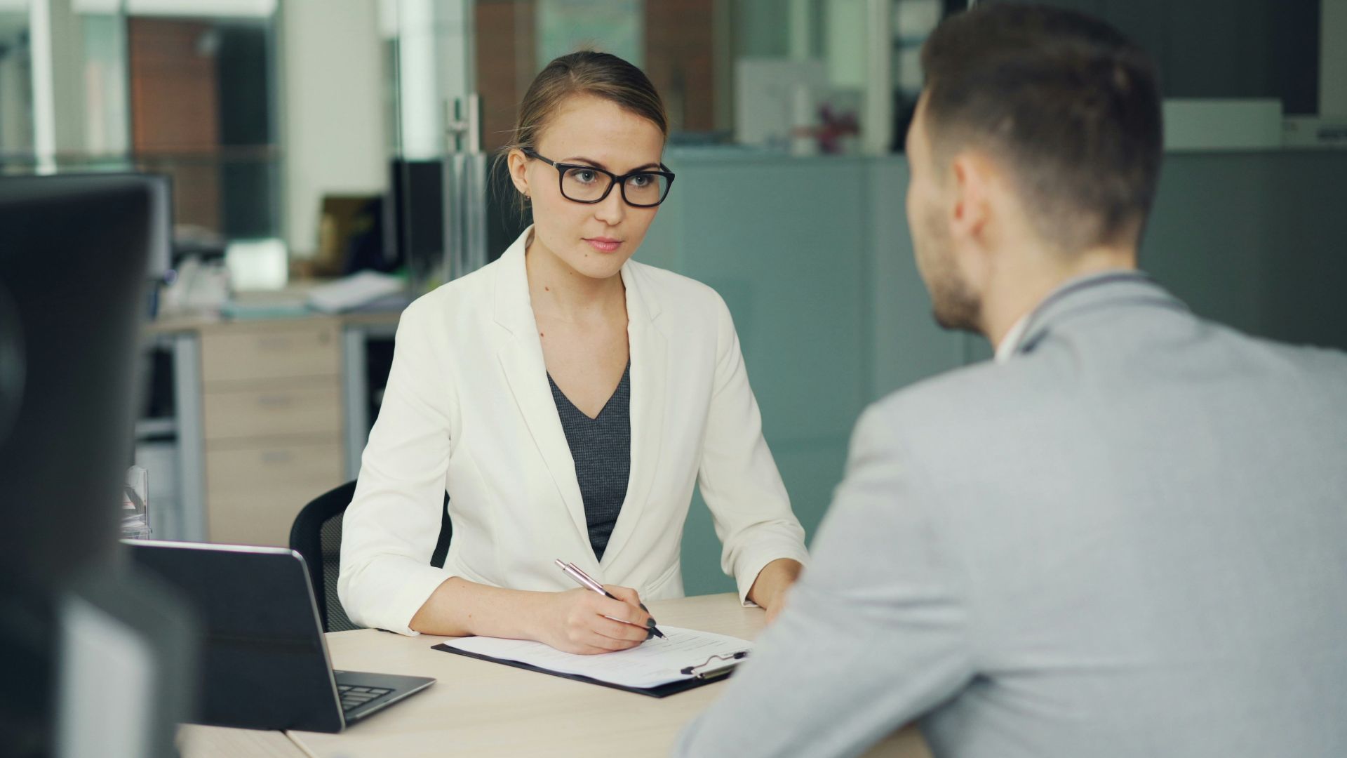 Woman in glasses interviews man at office desk.