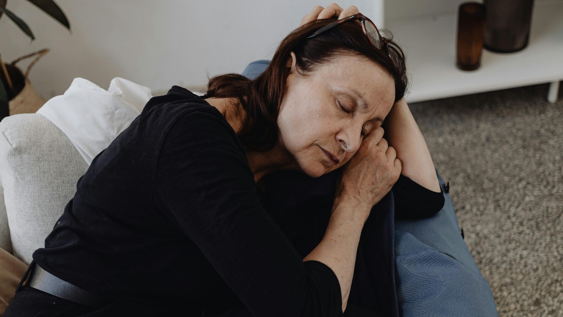 An elderly woman rests peacefully on a sofa in a cozy domestic setting, wearing eyeglasses.