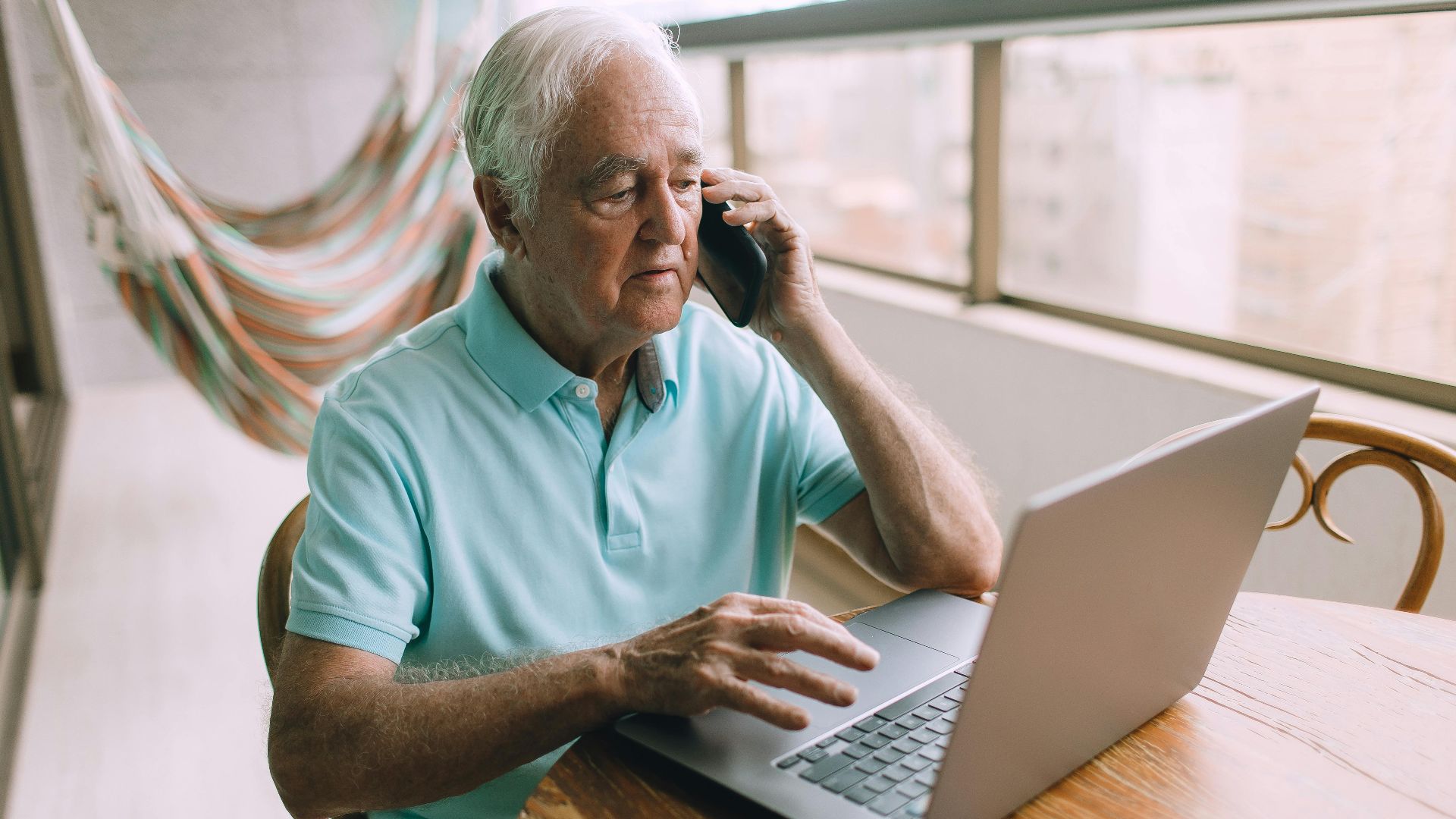 Elderly man multitasking with laptop and phone indoors, demonstrating technology use.