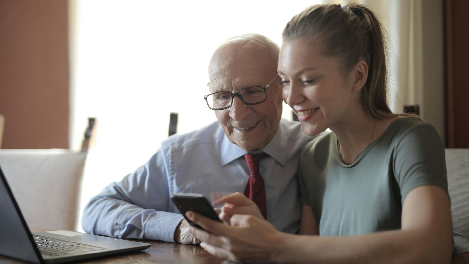 Smiling young woman in casual clothes showing smartphone to interested senior grandfather in formal shirt and eyeglasses while sitting at table near laptop
