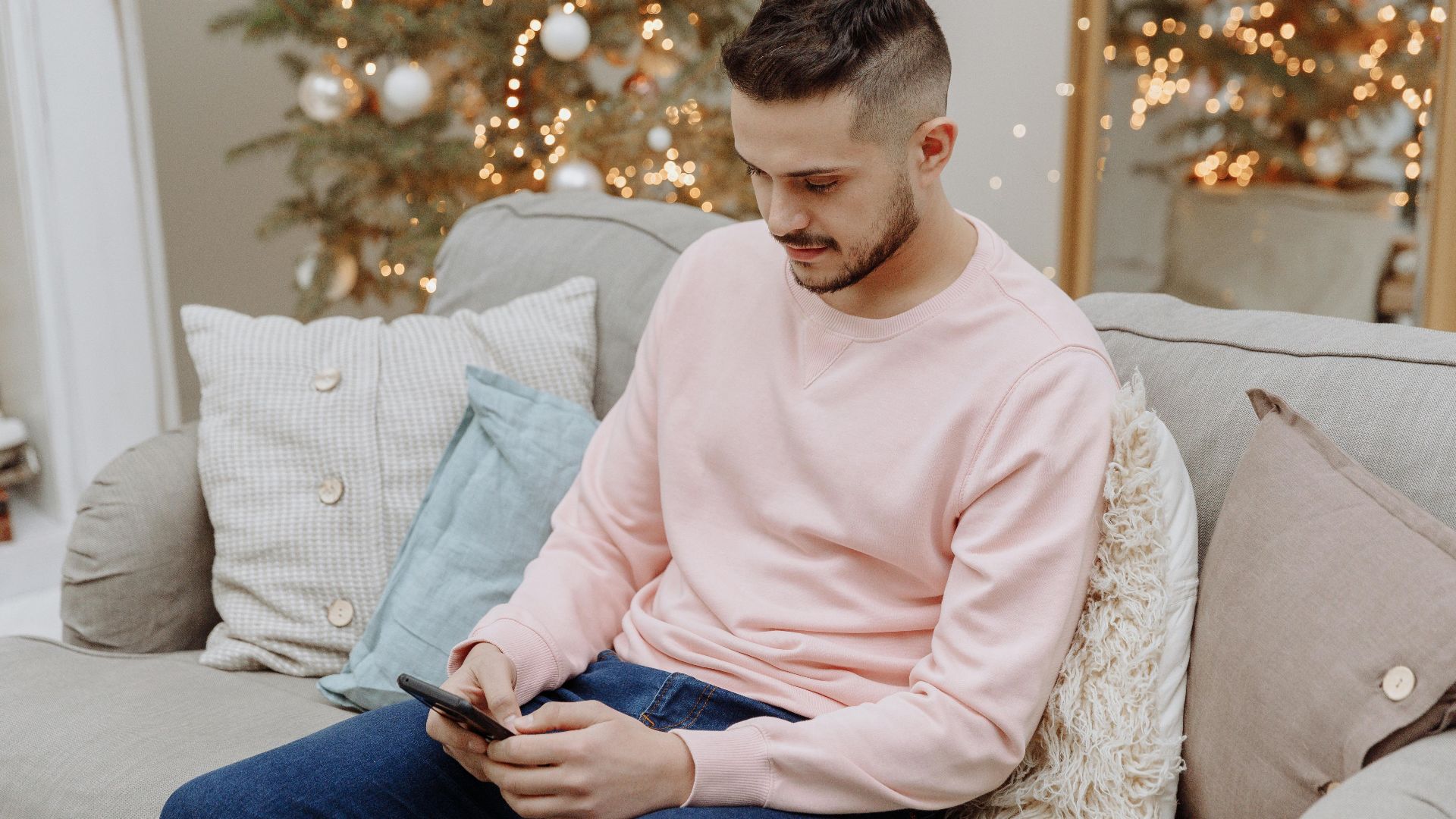 A man sitting on a sofa using a smartphone in a festive room with a Christmas tree.