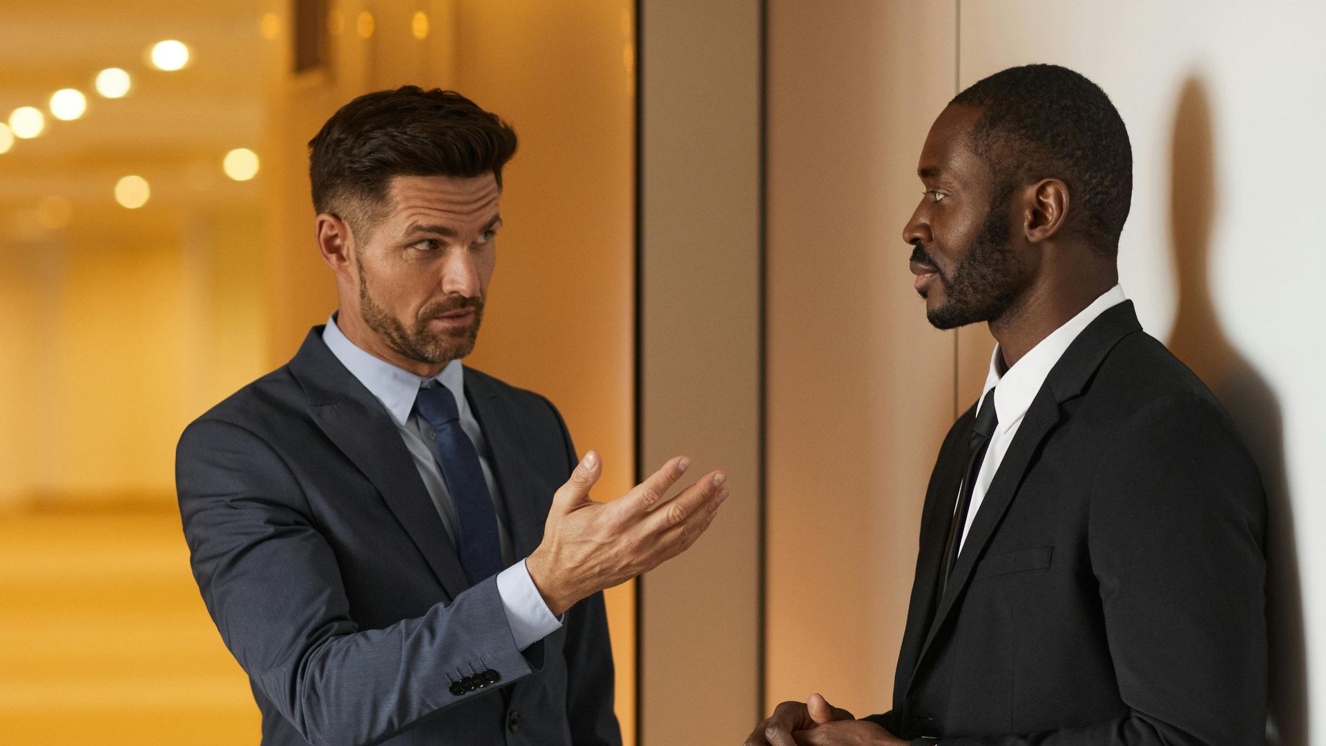 Two businessmen in formal attire engaged in a serious discussion in a modern office hallway.