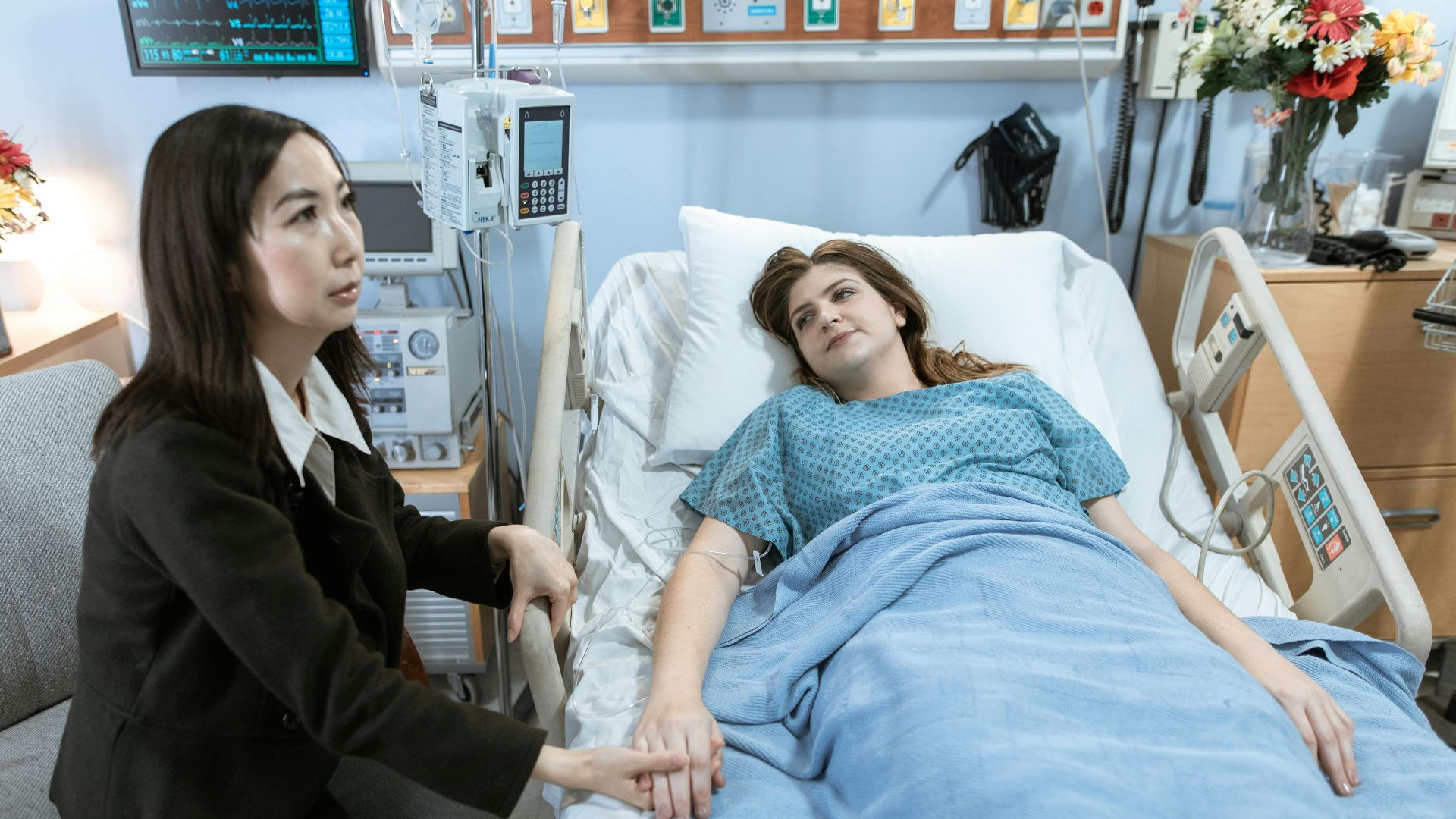 A woman visits a patient in a hospital room, holding hands for comfort.