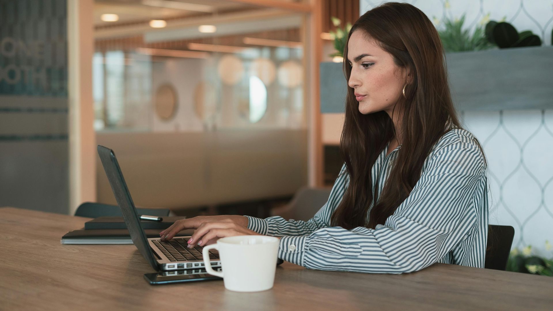 A professional woman working on her laptop at a modern office with a coffee cup nearby.