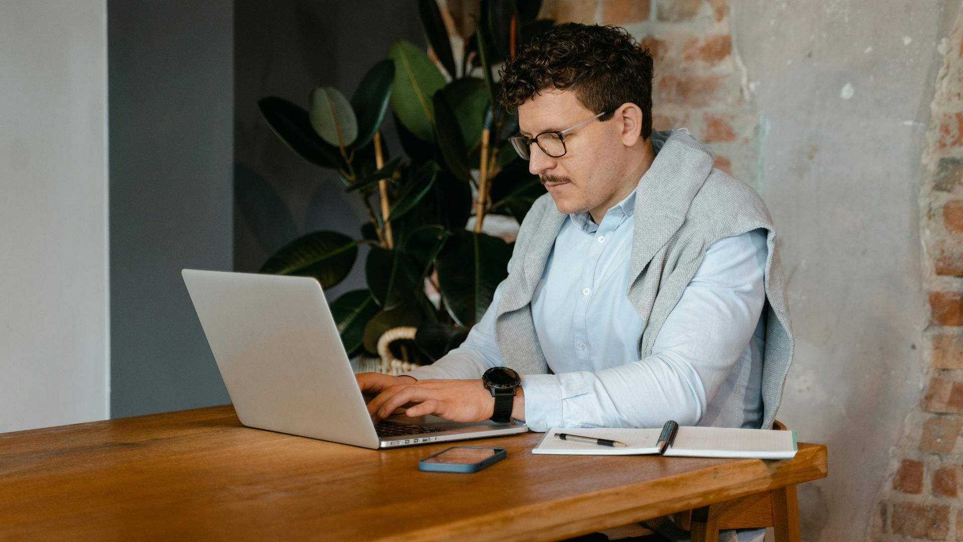Adult man using a laptop at a wooden table in a cozy workspace with exposed brick walls.