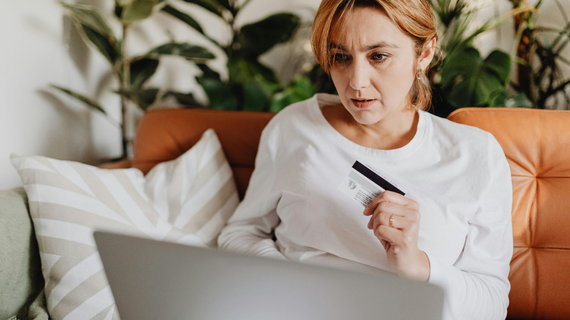 A woman sitting on a couch shopping online with a credit card and laptop in a cozy living room.