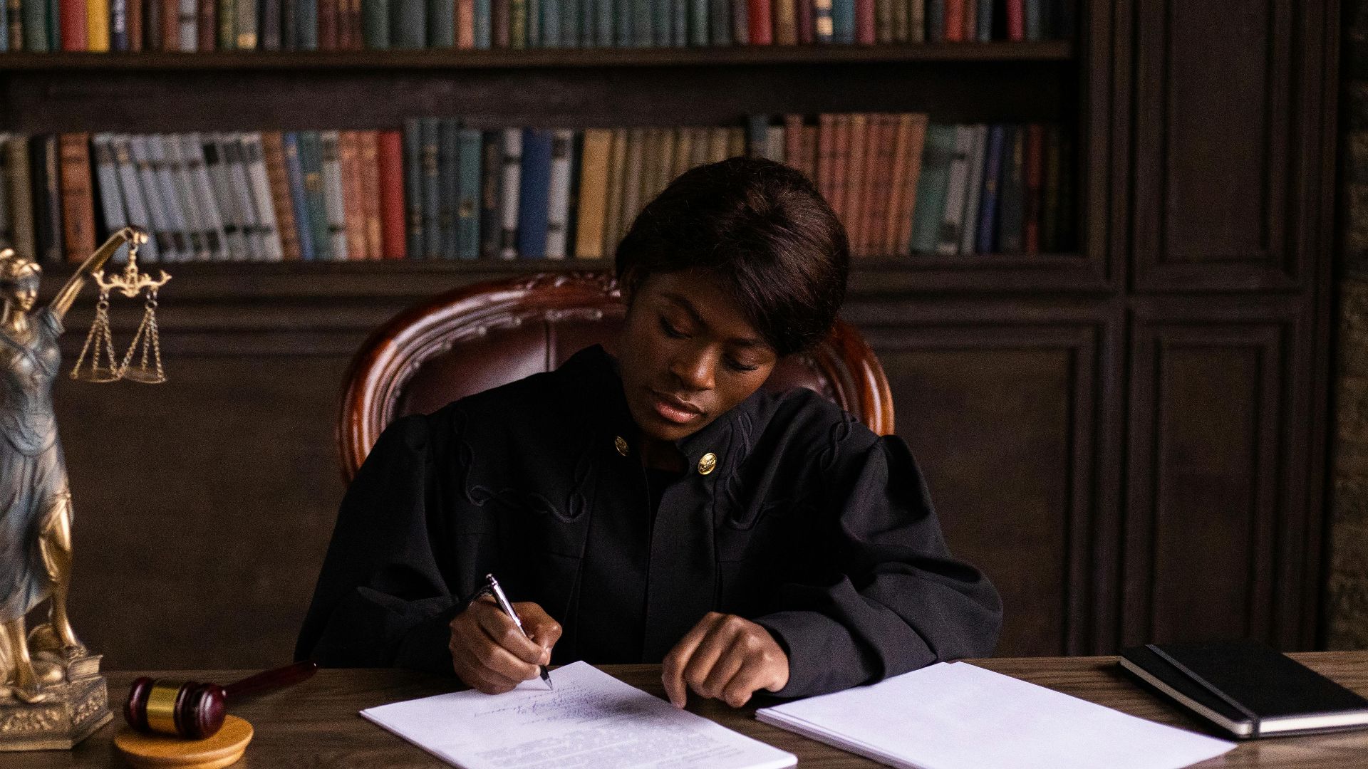 A judge in robes writing on a document at a desk in an office library with law books.