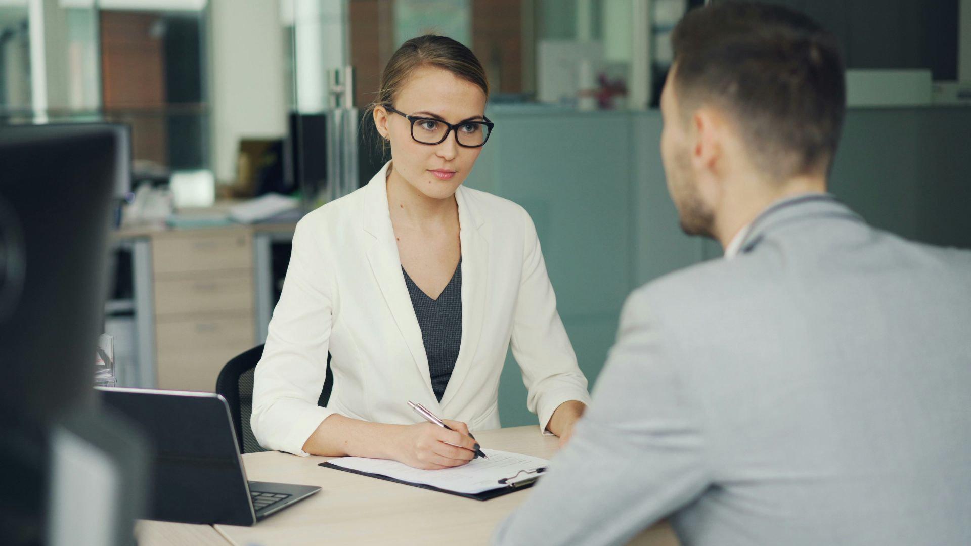 Woman in glasses interviews man at office desk.