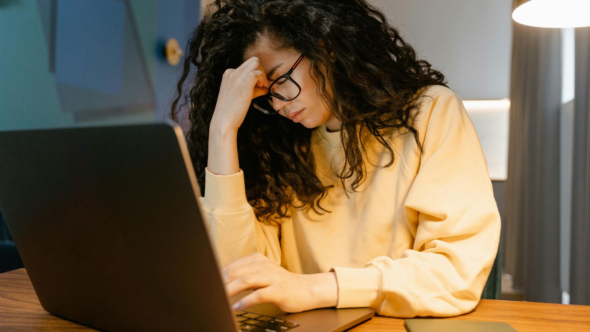 A tired woman with curly hair and glasses experiencing stress while working on a laptop.