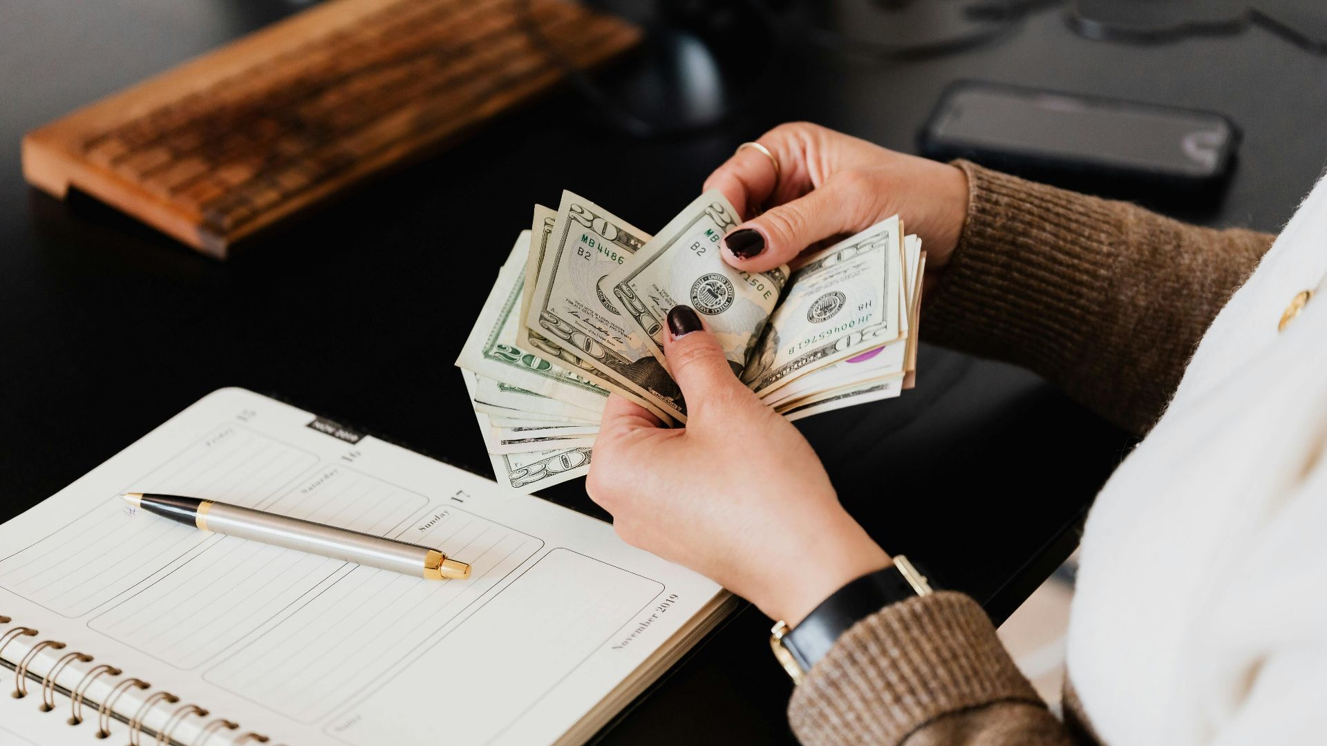 Unrecognizable elegant female in sweater counting dollar bills while sitting at wooden table with planner and pen