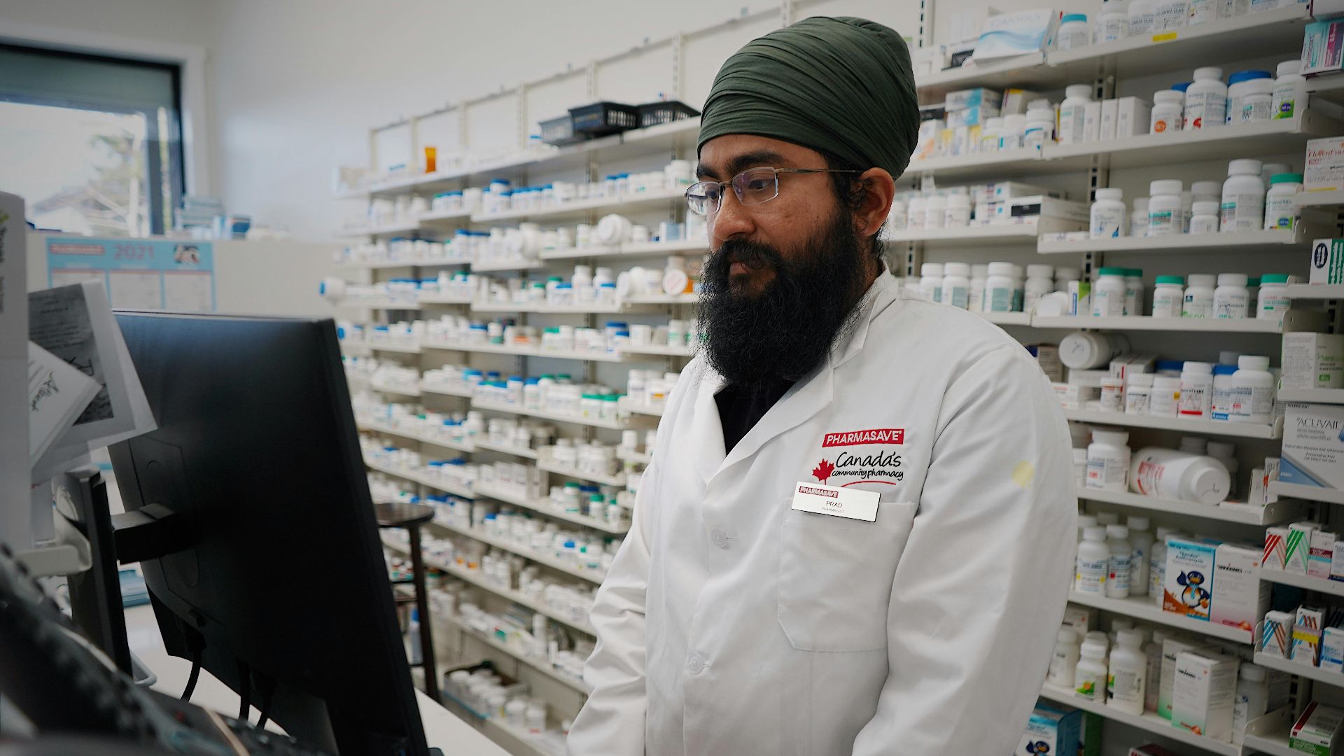 Pharmacist wearing a turban working at a computer in a pharmacy store with shelves filled with medicine.