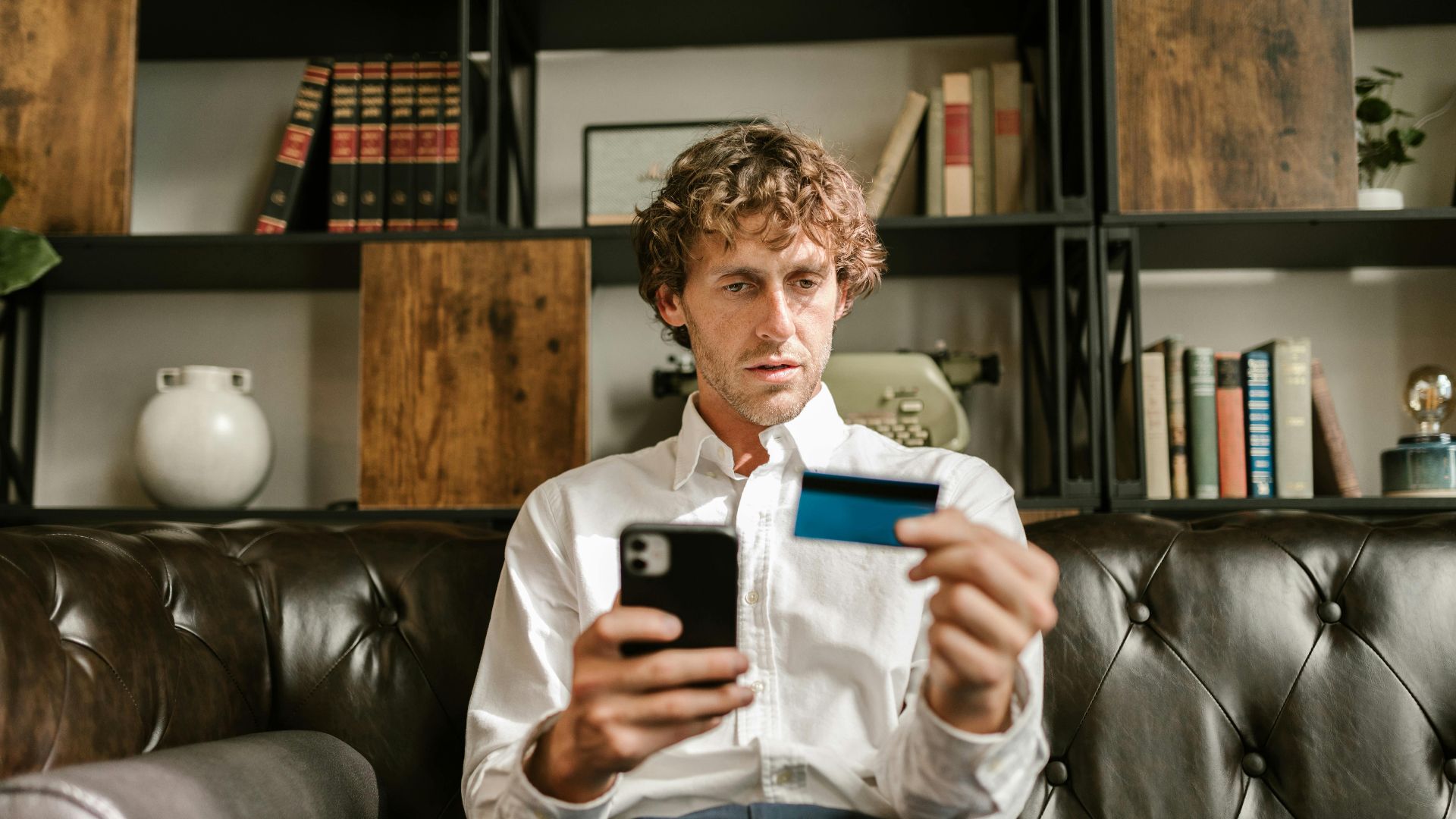 A man sitting on a leather sofa using a smartphone and credit card for online shopping.
