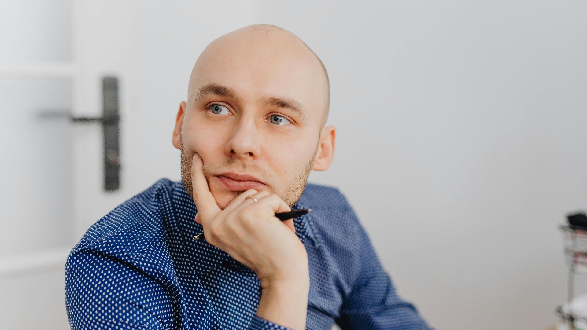 Bald man in blue shirt thoughtfully holding a pen indoors, gazing into distance.