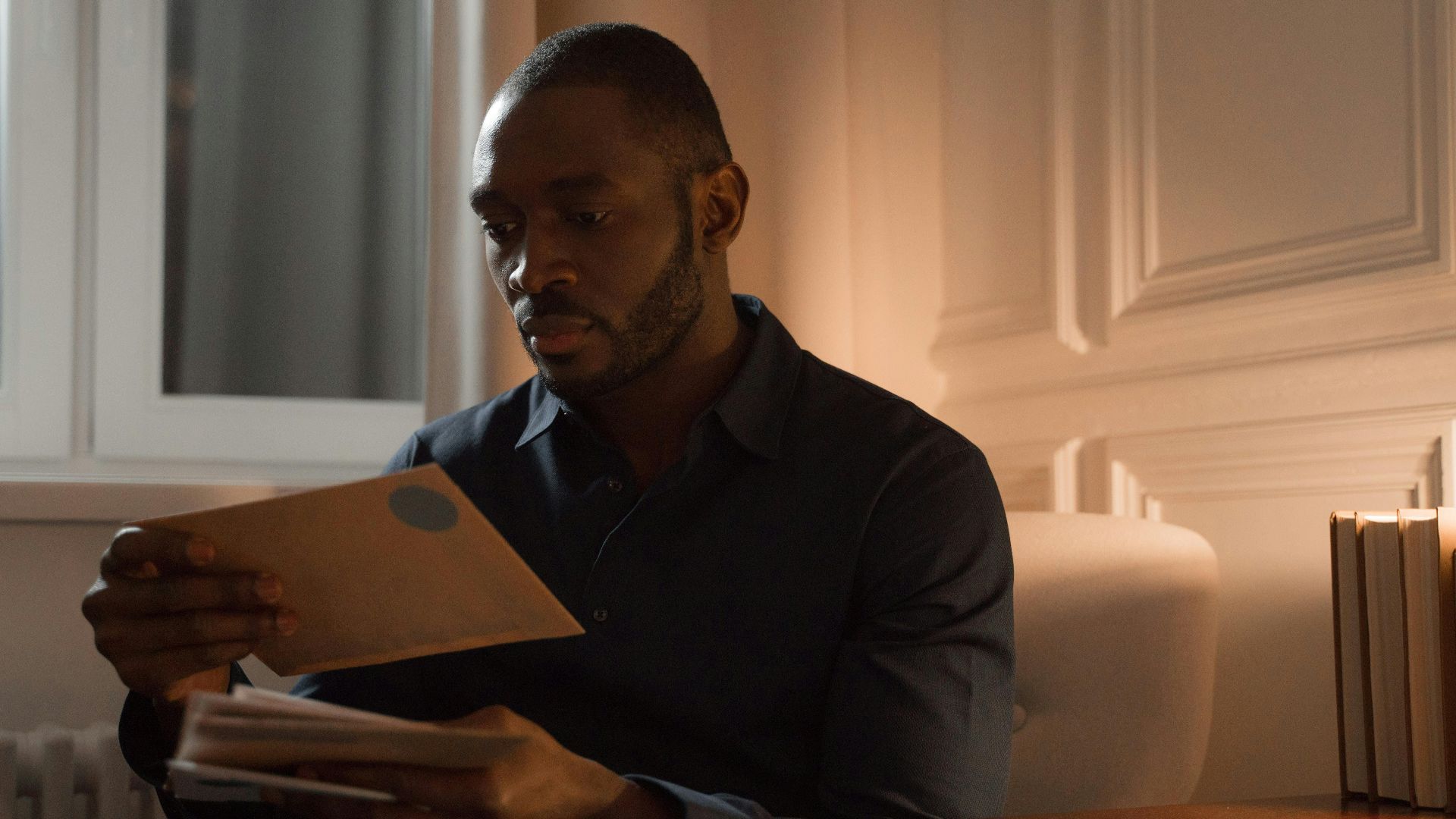 A man with a mustache reads letters while sitting in a warmly lit home interior.