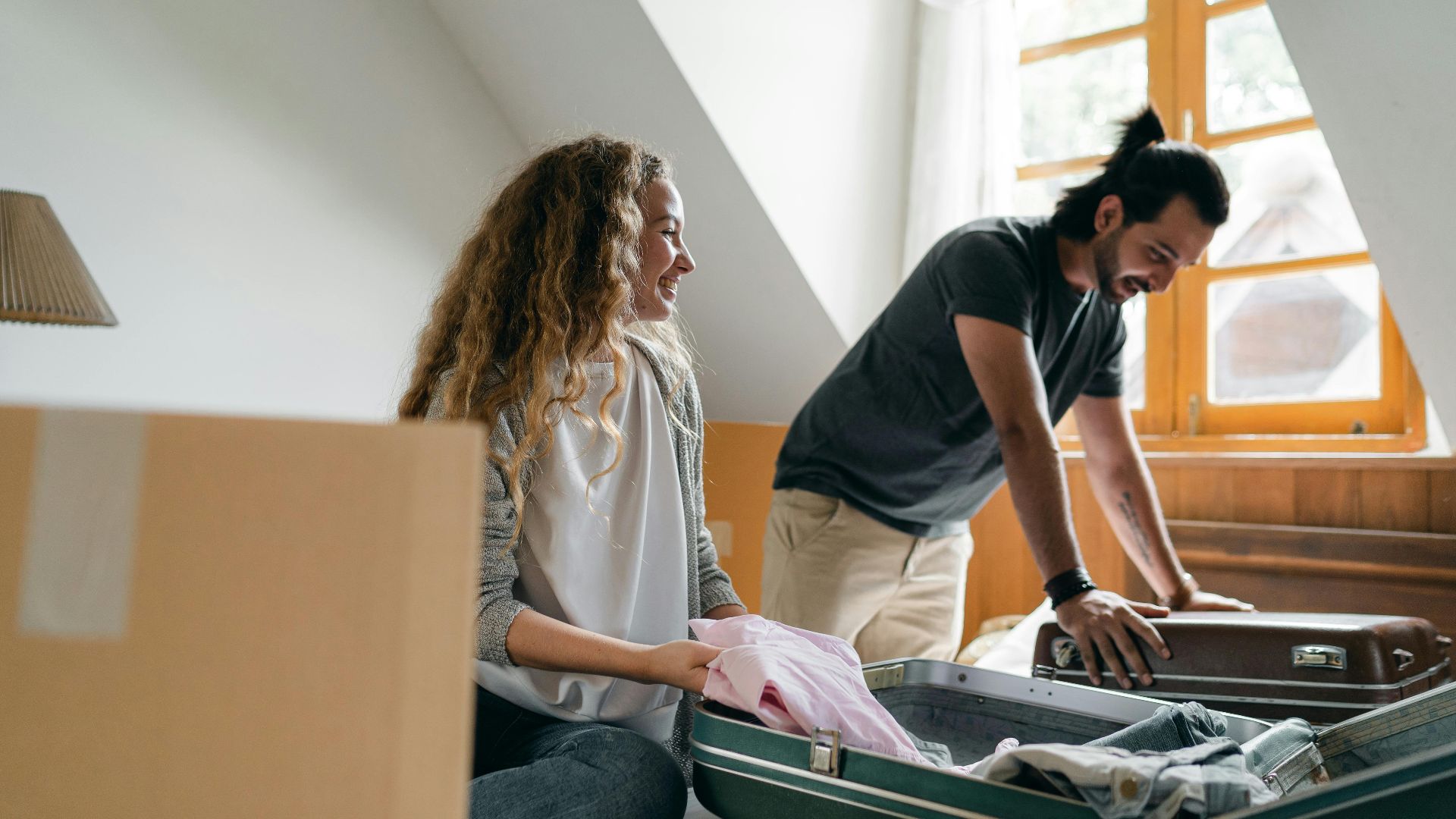 Positive young couple smiling and packing suitcases together in empty bedroom while preparing to move into new house