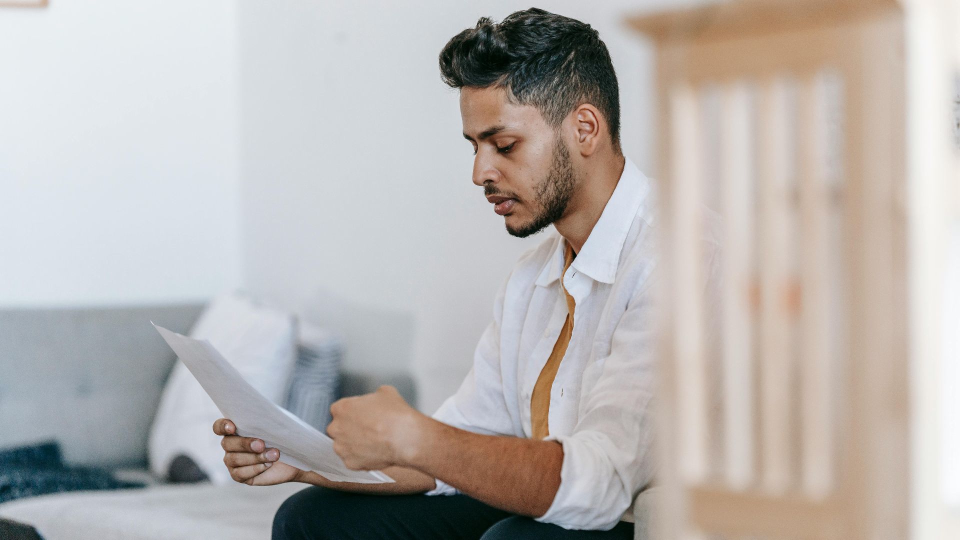 Concentrated young ethnic male wearing casual outfit reading documents and sitting on couch in modern living room