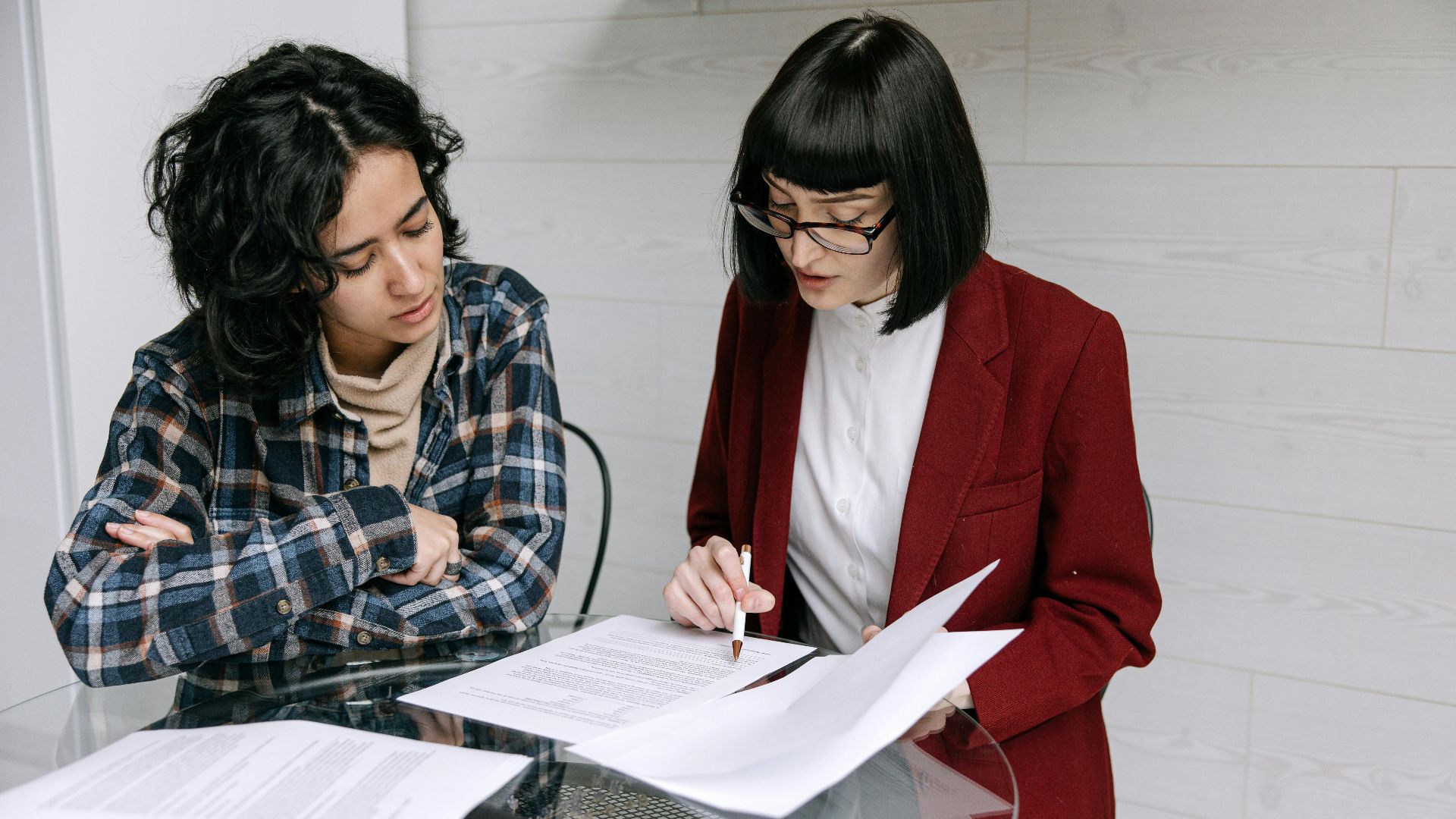Two women collaborating at a glass table, reviewing business documents in a modern office setting.