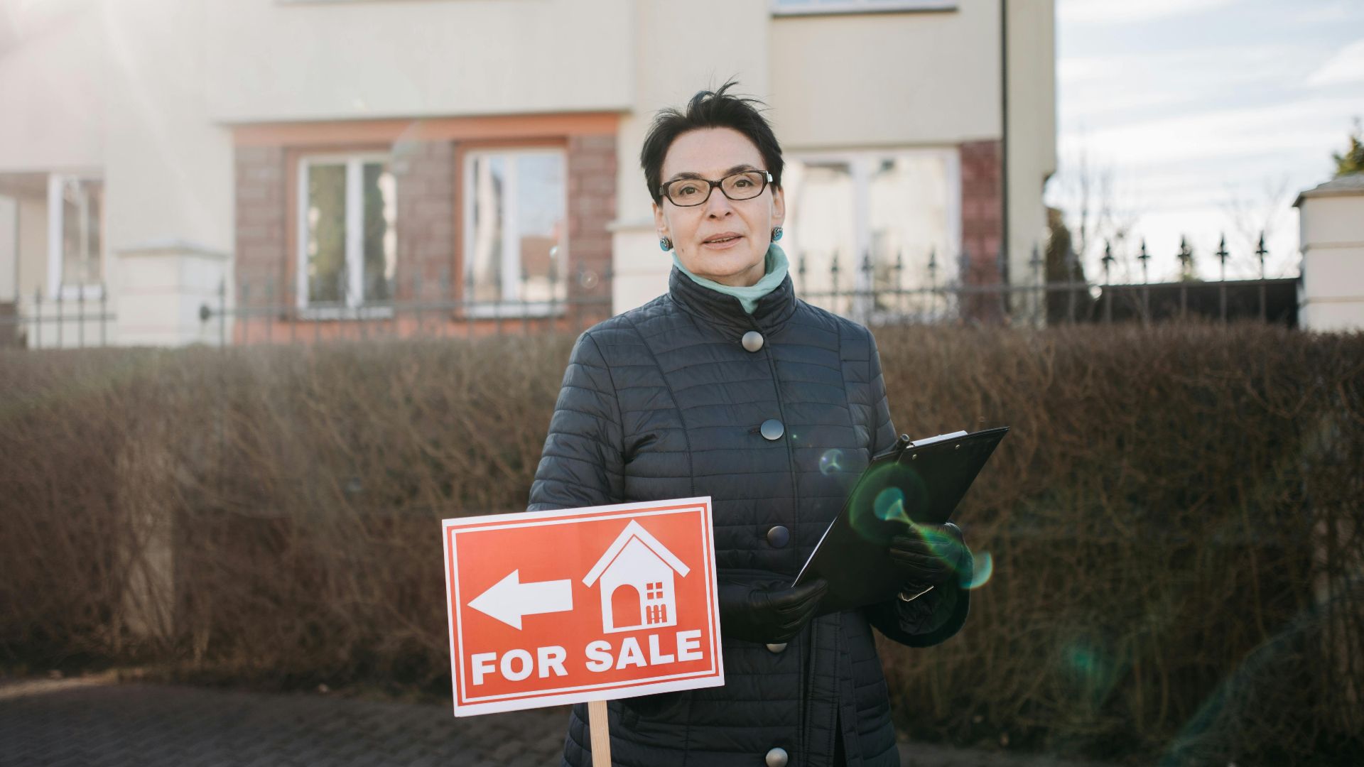 Woman realtor with eyeglasses holding a clipboard and 'For Sale' sign outside a house.