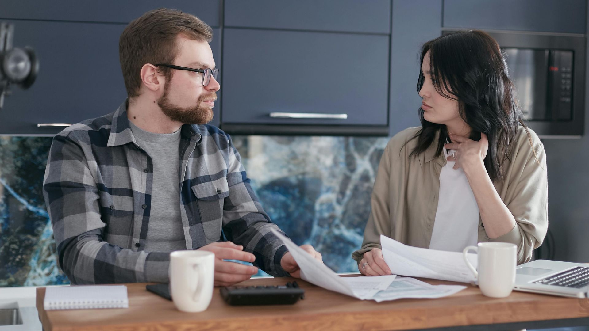 A young couple sitting at a table discussing bills and financial plans in a modern kitchen.