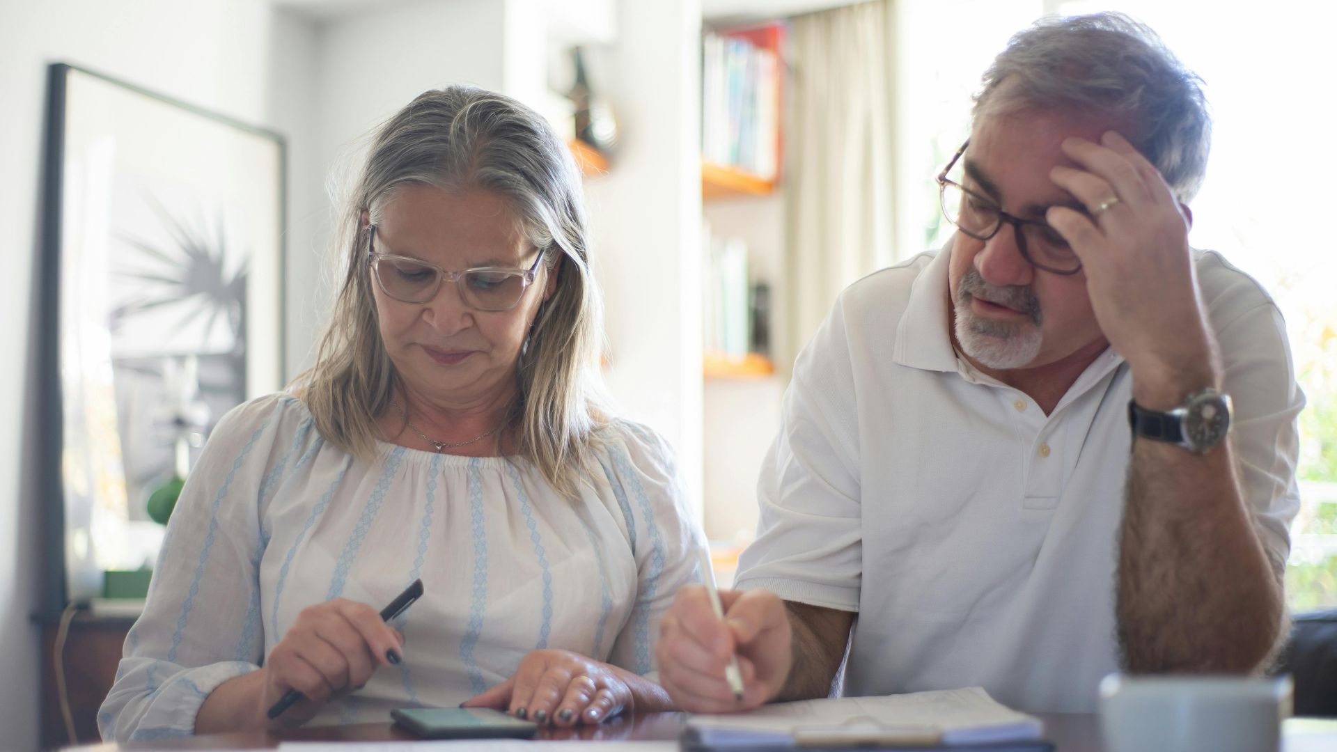 Senior couple calculating expenses at home office desk with documents and notes.