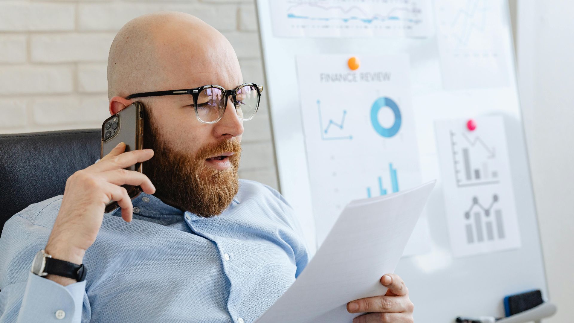 Bald man with beard holding smartphone and papers, reviewing financial charts in office.