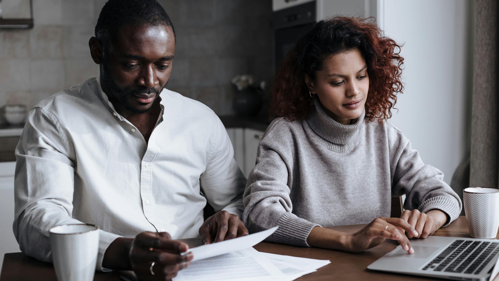 A diverse couple collaborates on a laptop at their home, handling documents over coffee.