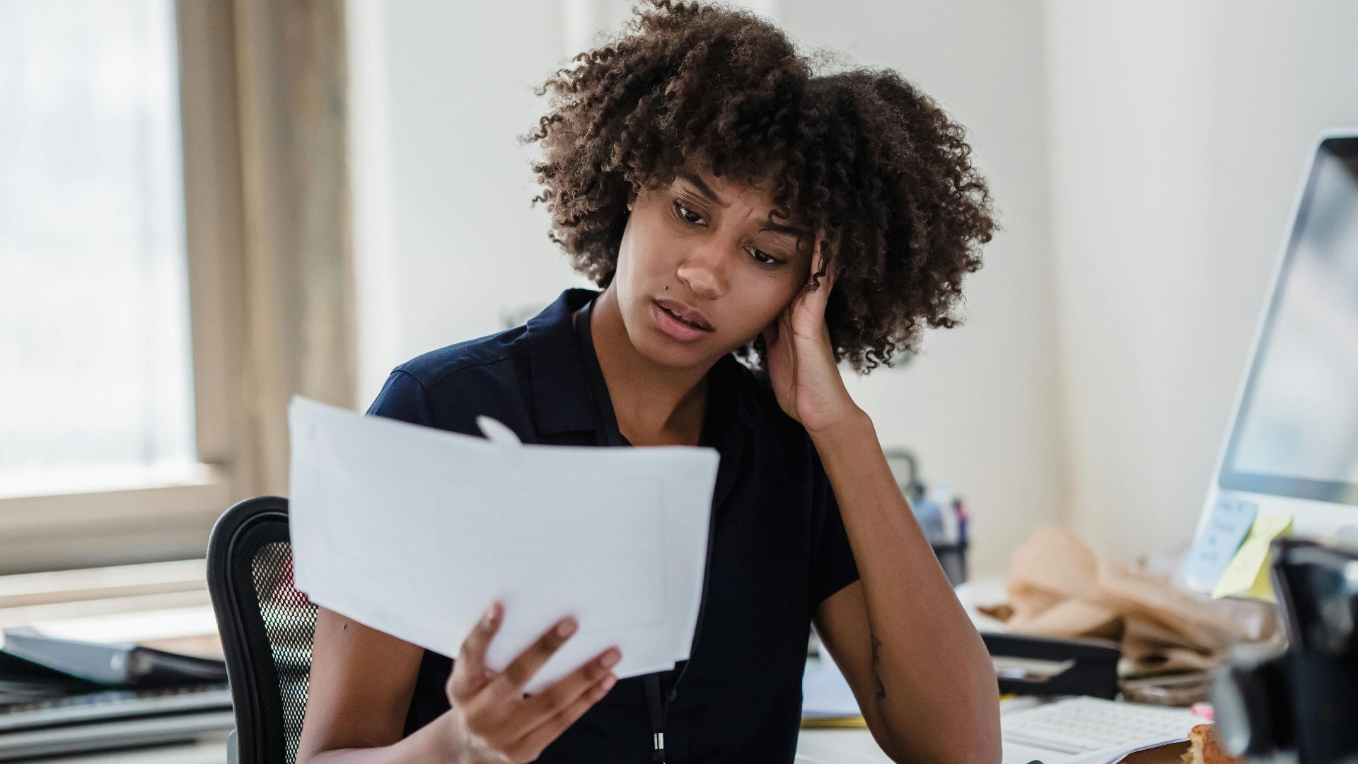 A professional woman with curly hair reviews documents at her office desk, deep in thought.