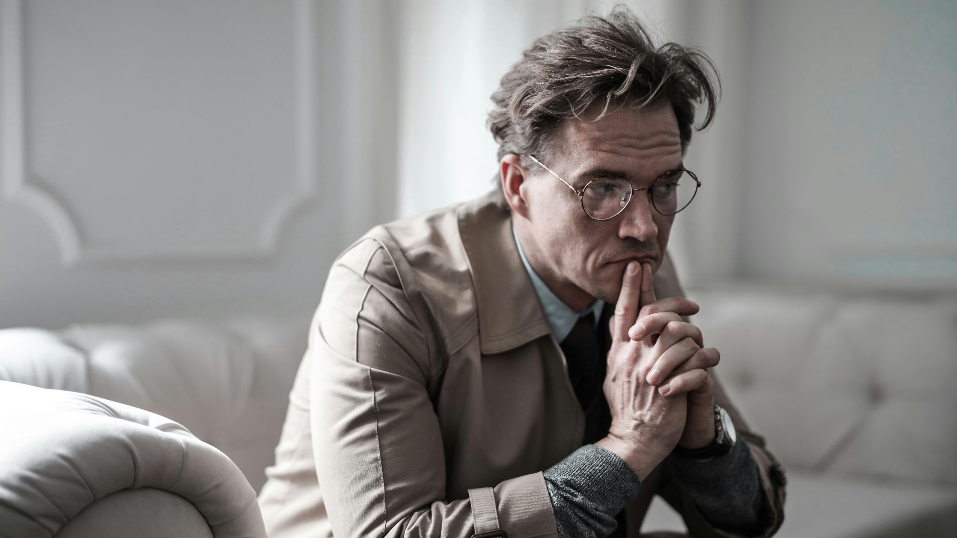 A thoughtful man wearing glasses sits on a sofa in a classic interior, lost in deep contemplation.