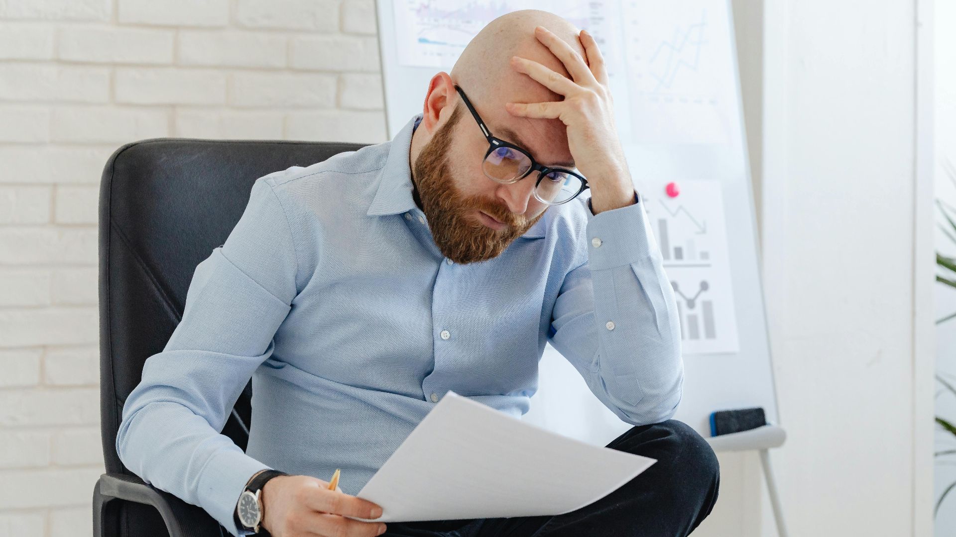 Bald man with glasses sitting in office looking worried at document.