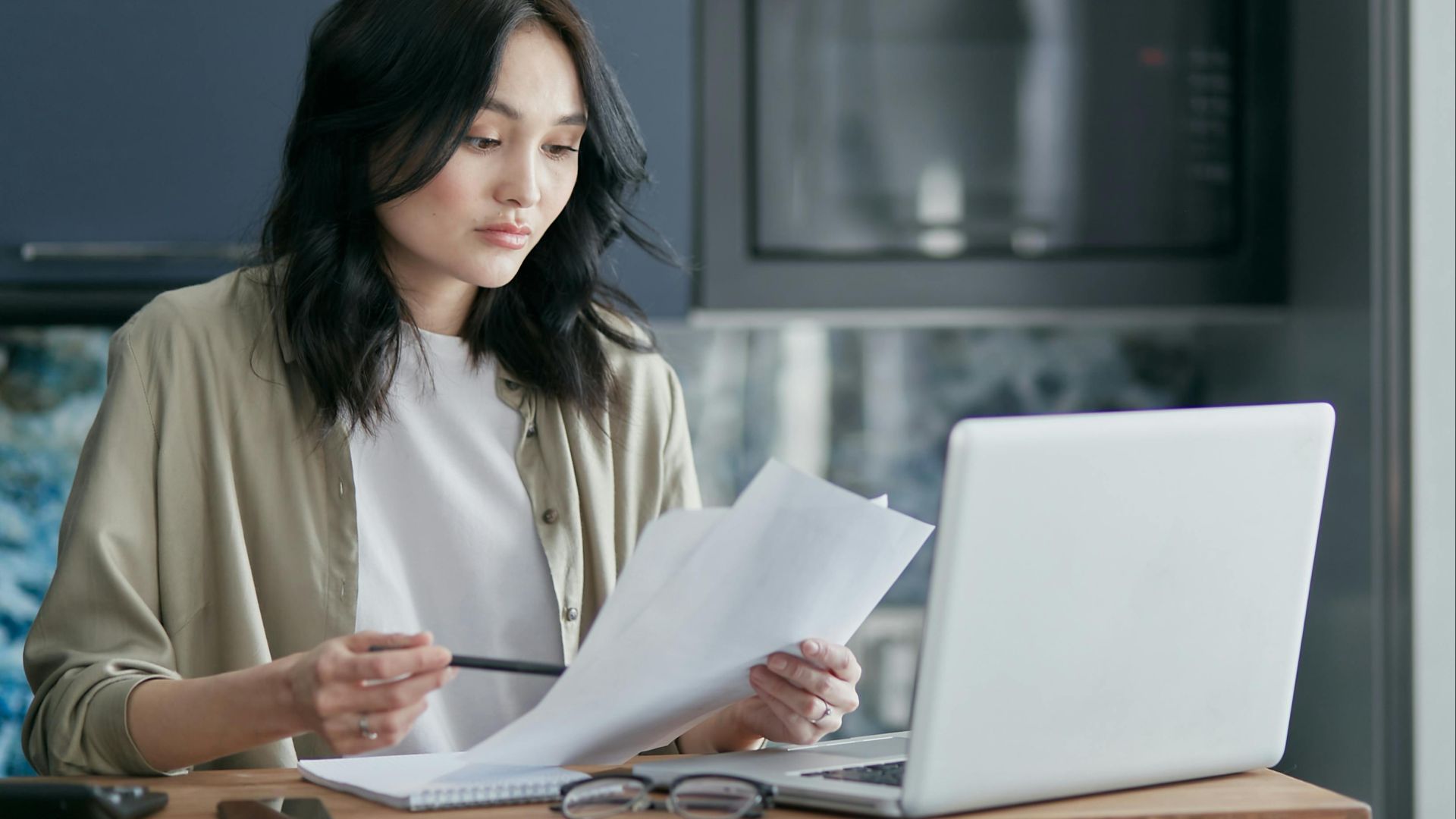 Concentrated woman reviewing documents and working on a laptop at home office setup in kitchen.