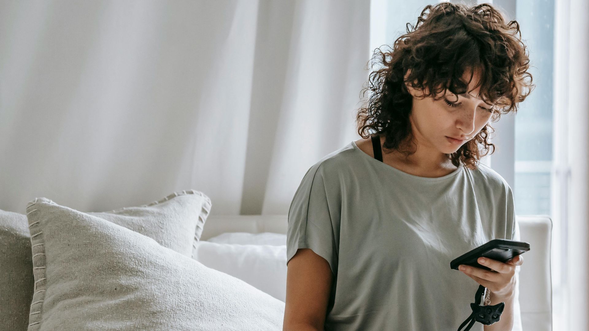Concentrated young ethnic woman with curly hair in casual clothes messaging on mobile phone and stroking cat while relaxing on comfortable bed during weekend
