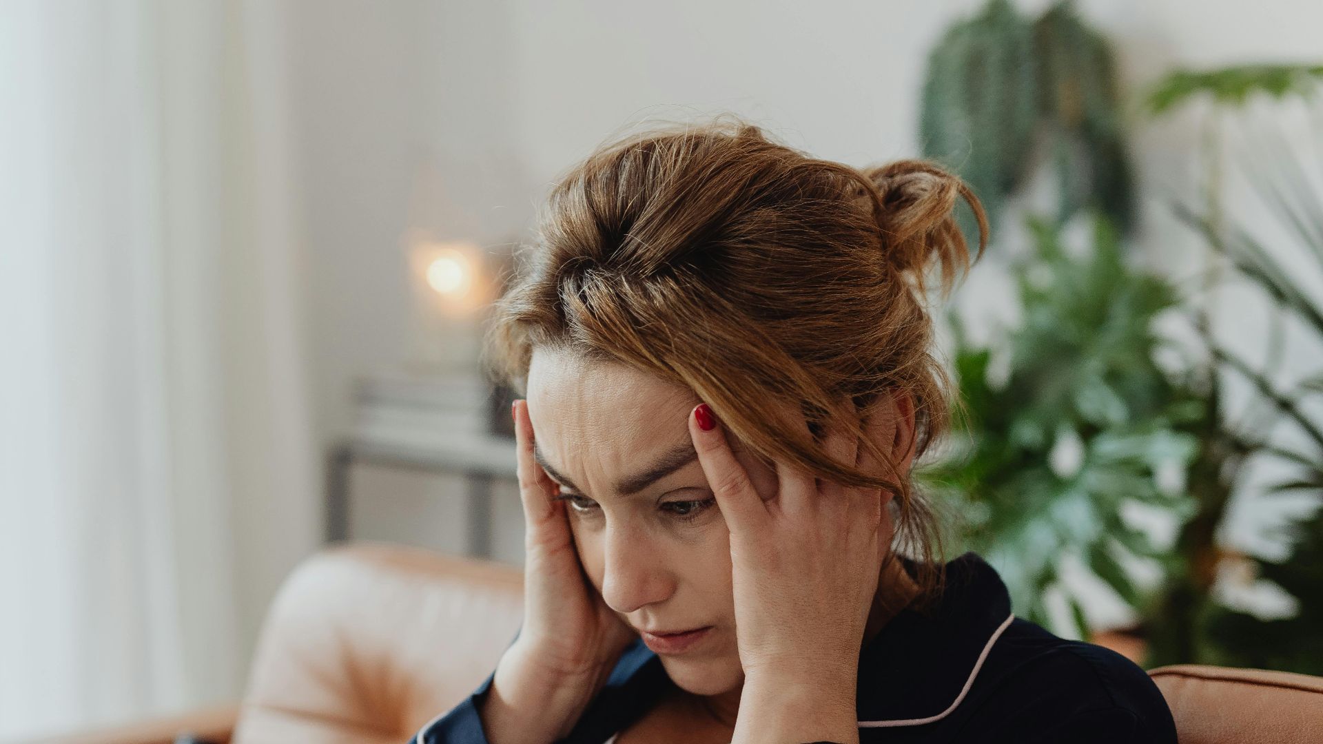 A woman in pajamas sits indoors with hands on head, showing stress and frustration. Perfect for mental health themes.