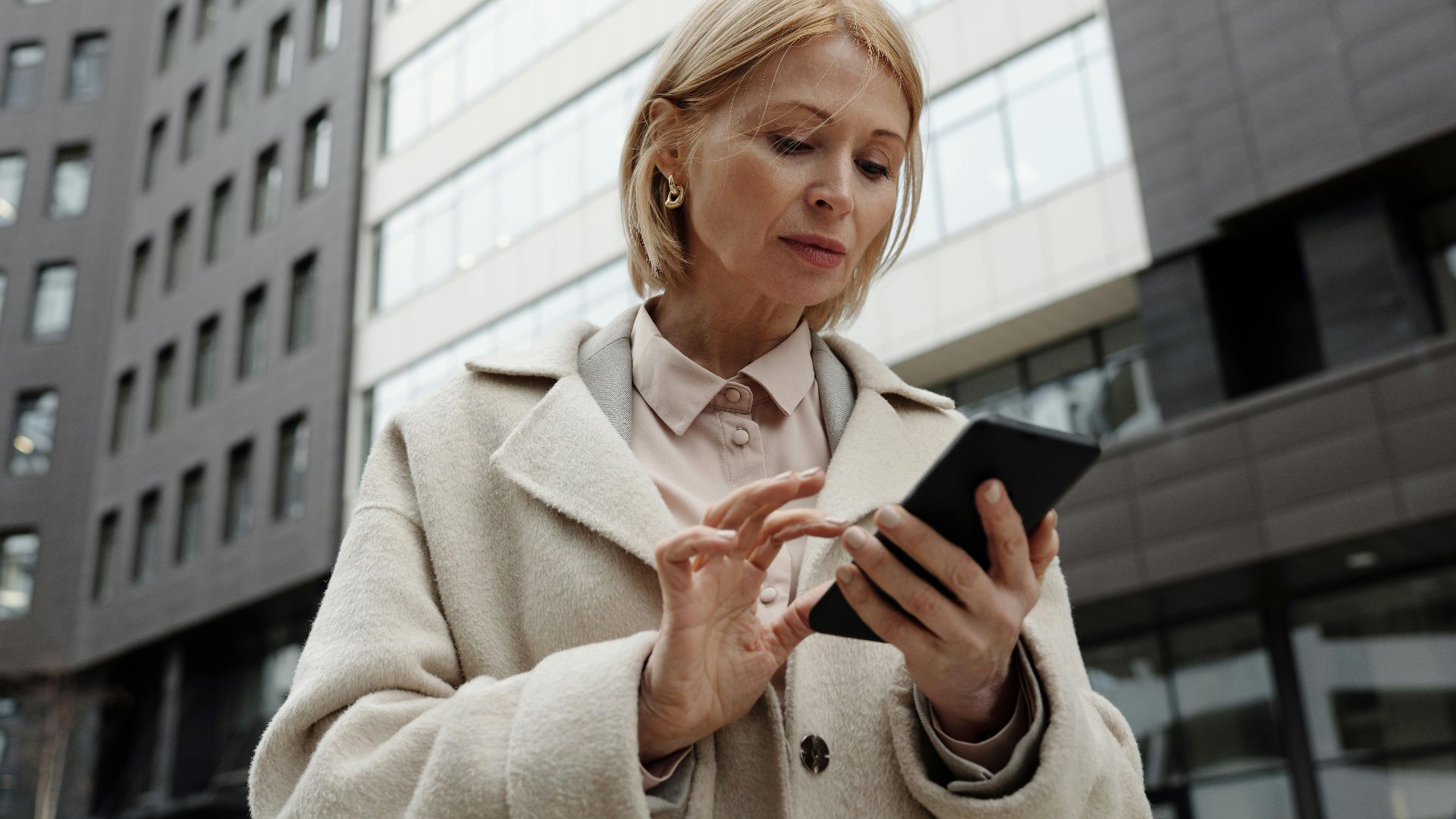 Woman in coat using smartphone in front of modern building. Professional and focused expression.