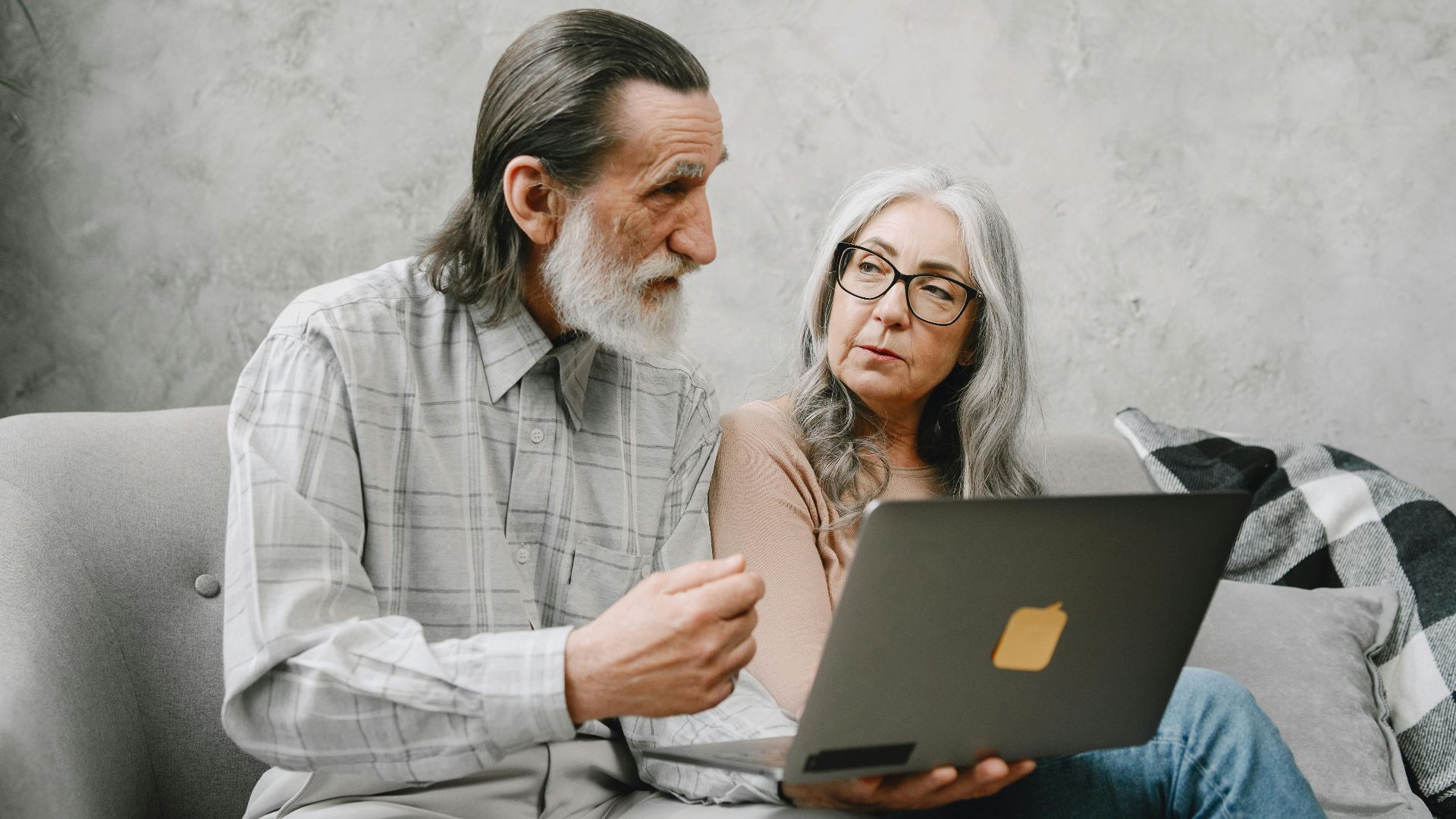 Elderly couple engaged in discussion while using a laptop at home.