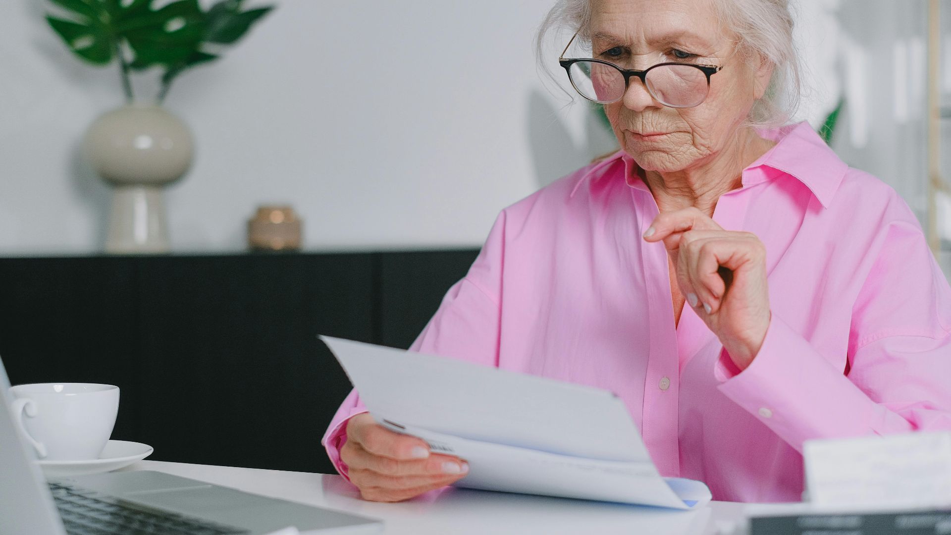 Elderly woman in pink shirt reading documents at home workspace.