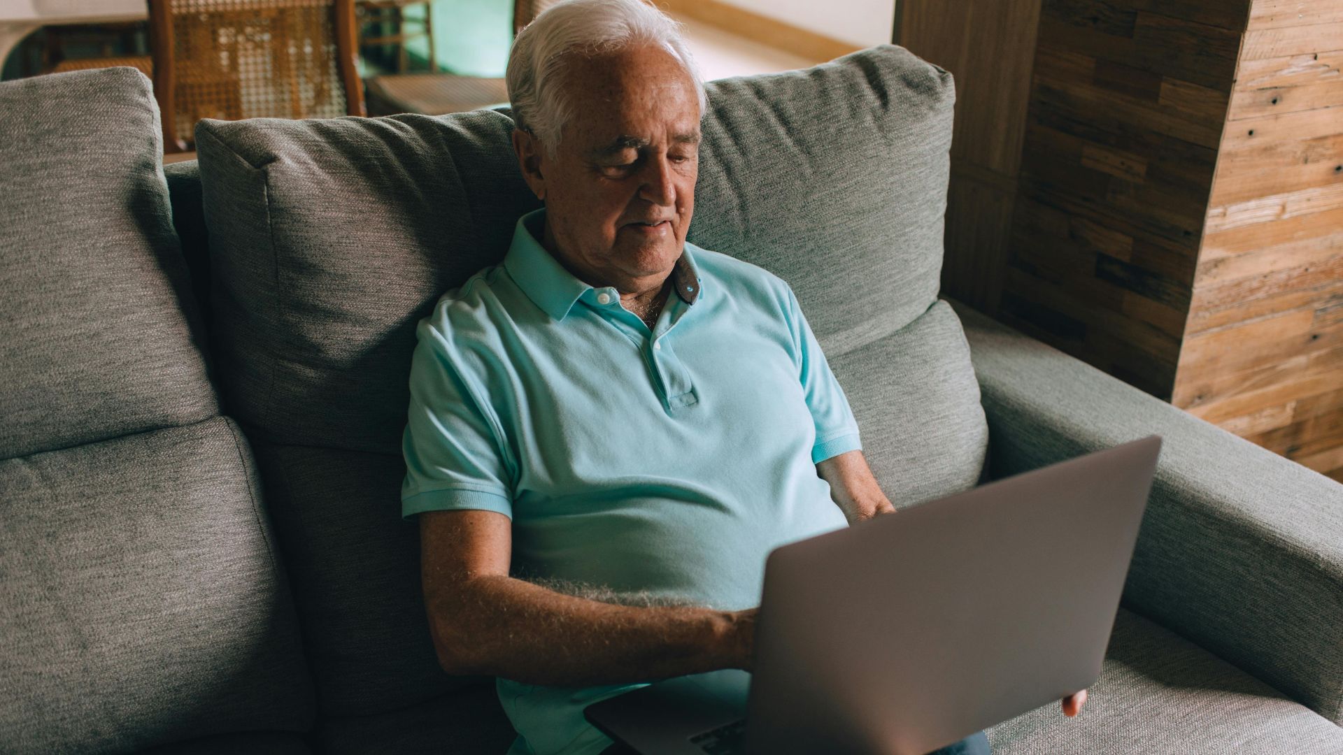 Senior man in blue polo shirt using a laptop while sitting on a cozy couch indoors.