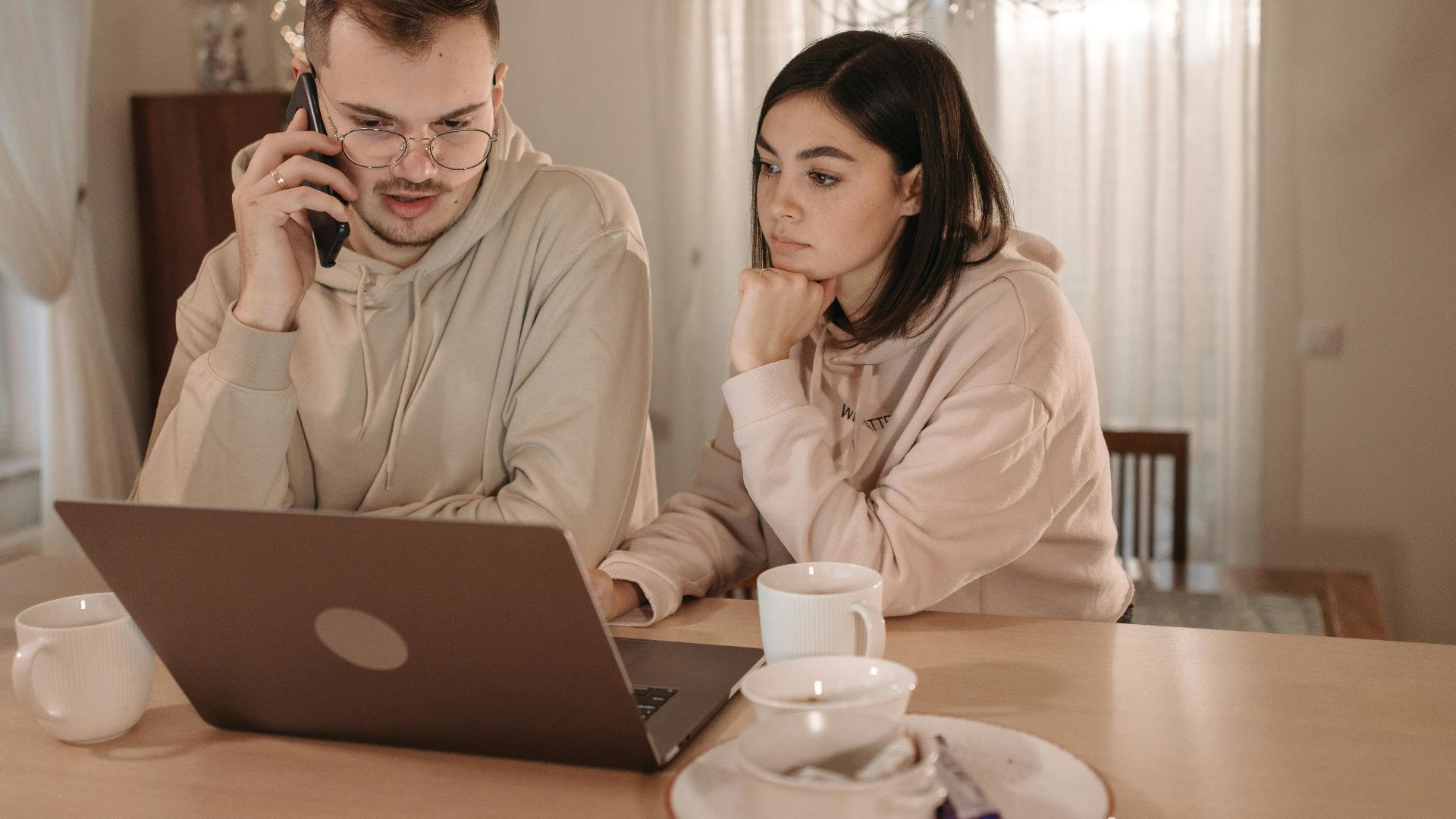 A couple in hoodies focused on work using a laptop and smartphone at home.