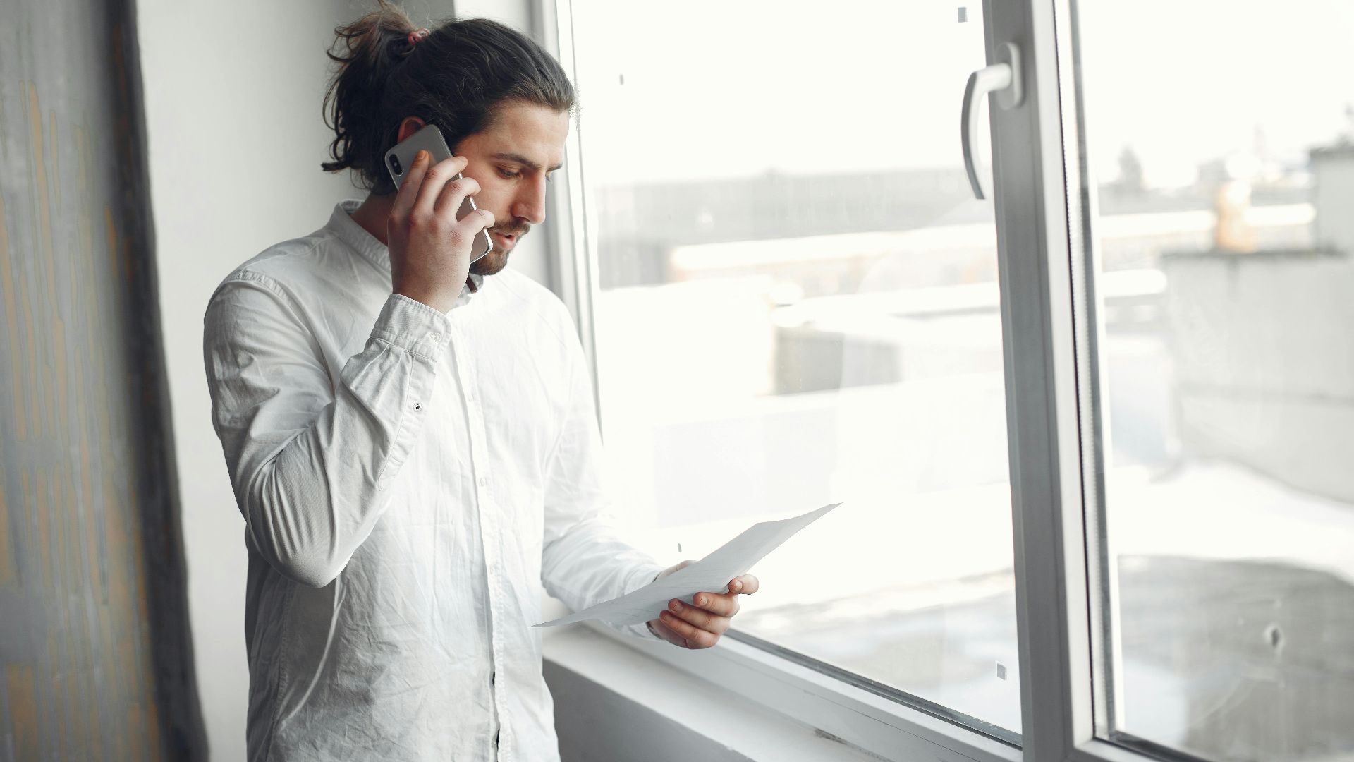 Young man in white shirt, on phone call holding a document, standing by a large window.