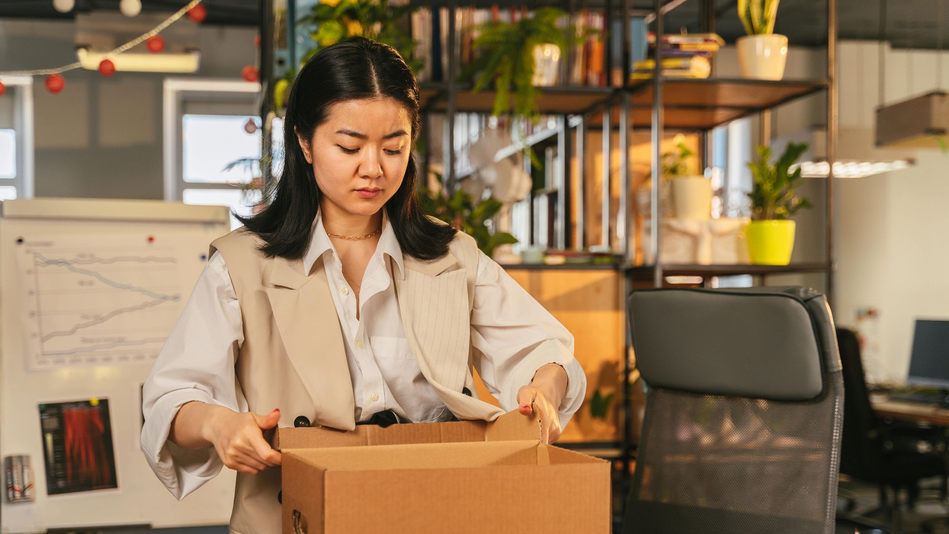An Asian woman packs a box labeled 'FIRED', symbolizing job loss in an office setting.