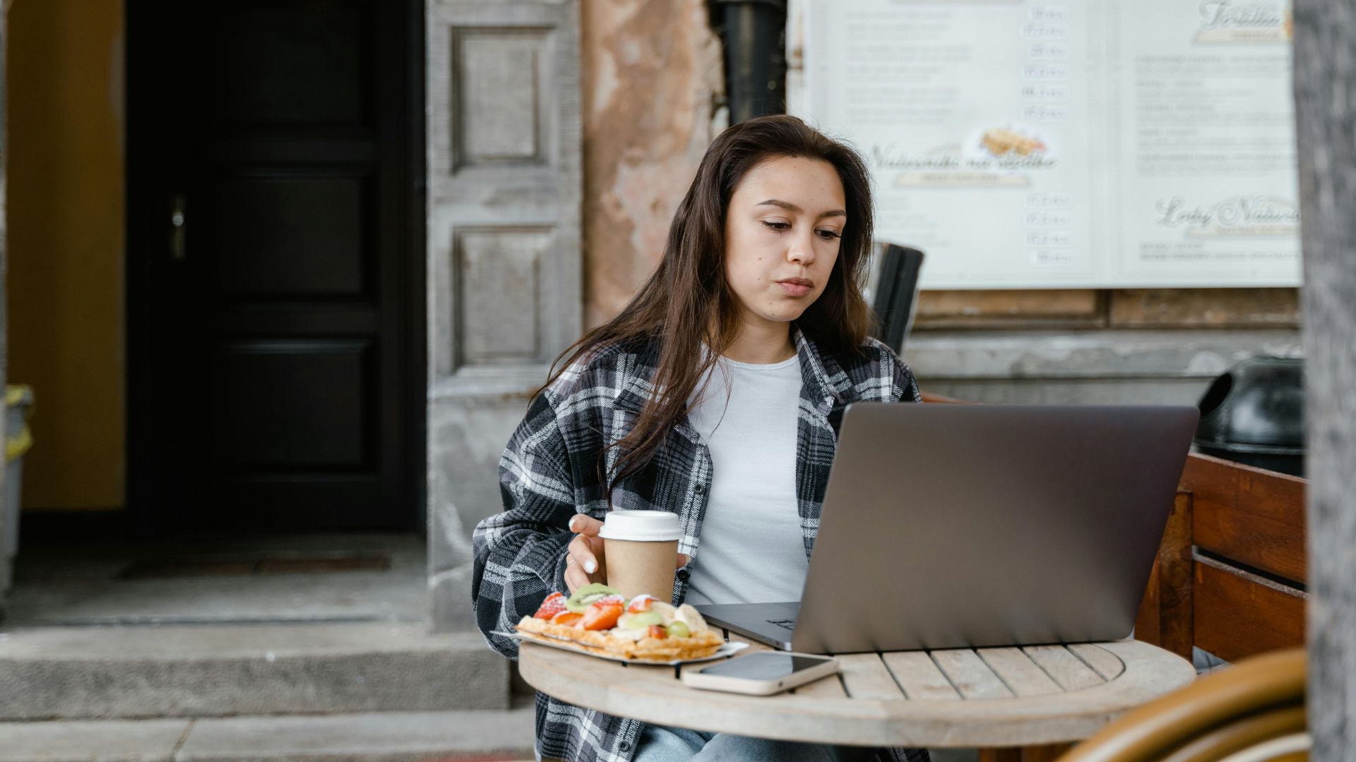 Woman working remotely on a laptop, sitting at an outdoor café with coffee and snacks.