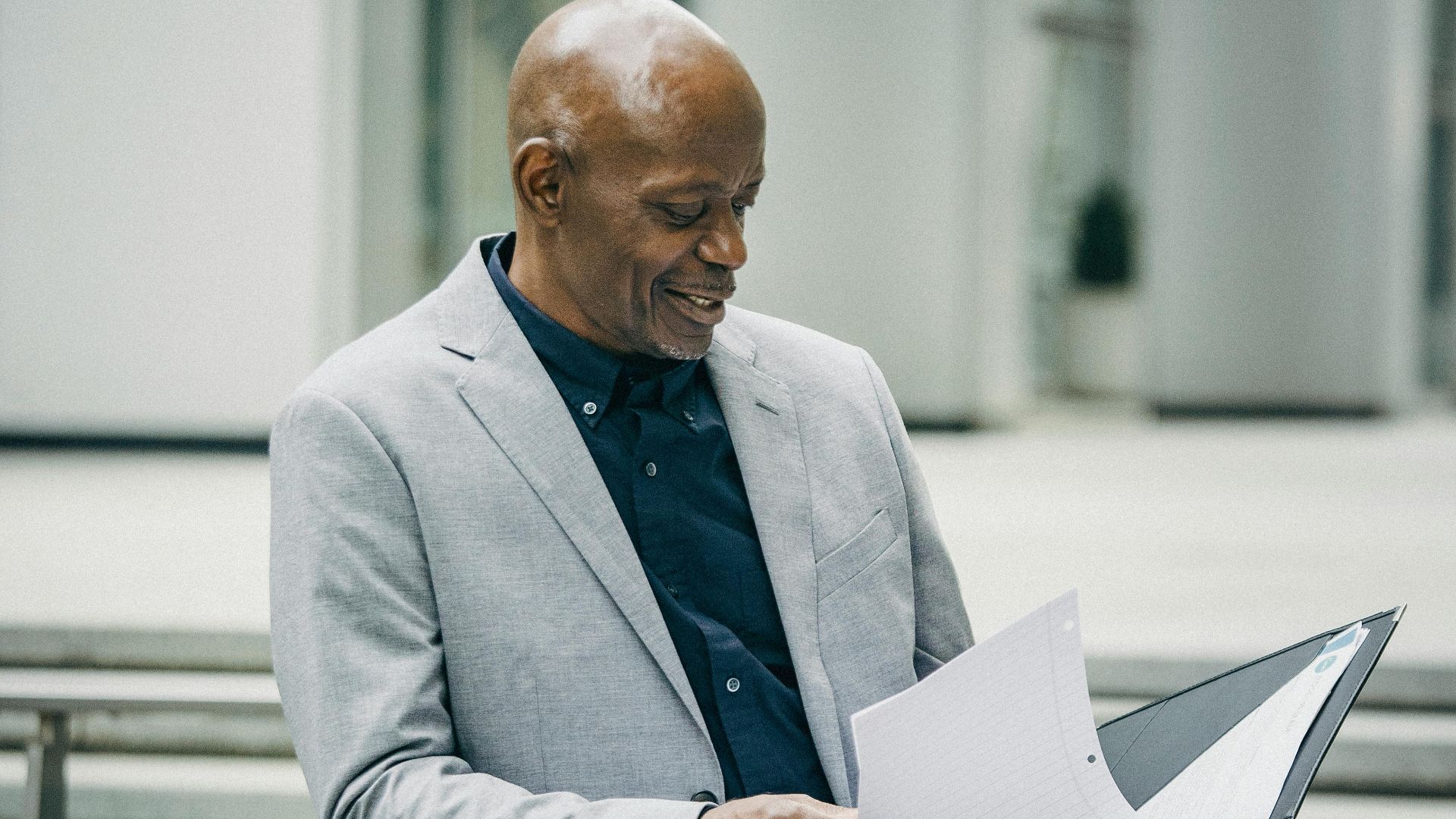Content middle aged African American male manager in formal suit smiling while checking documents standing near modern building