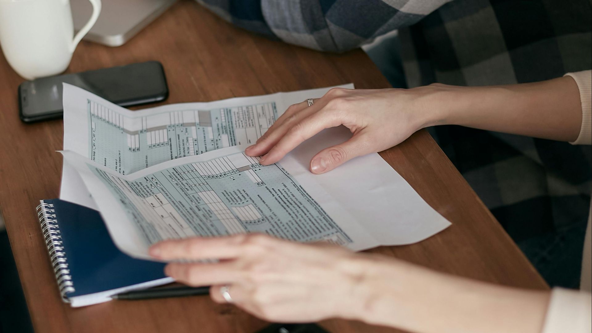 Close-up of hands organizing tax forms on a desk with a calculator, laptop, and notebook.