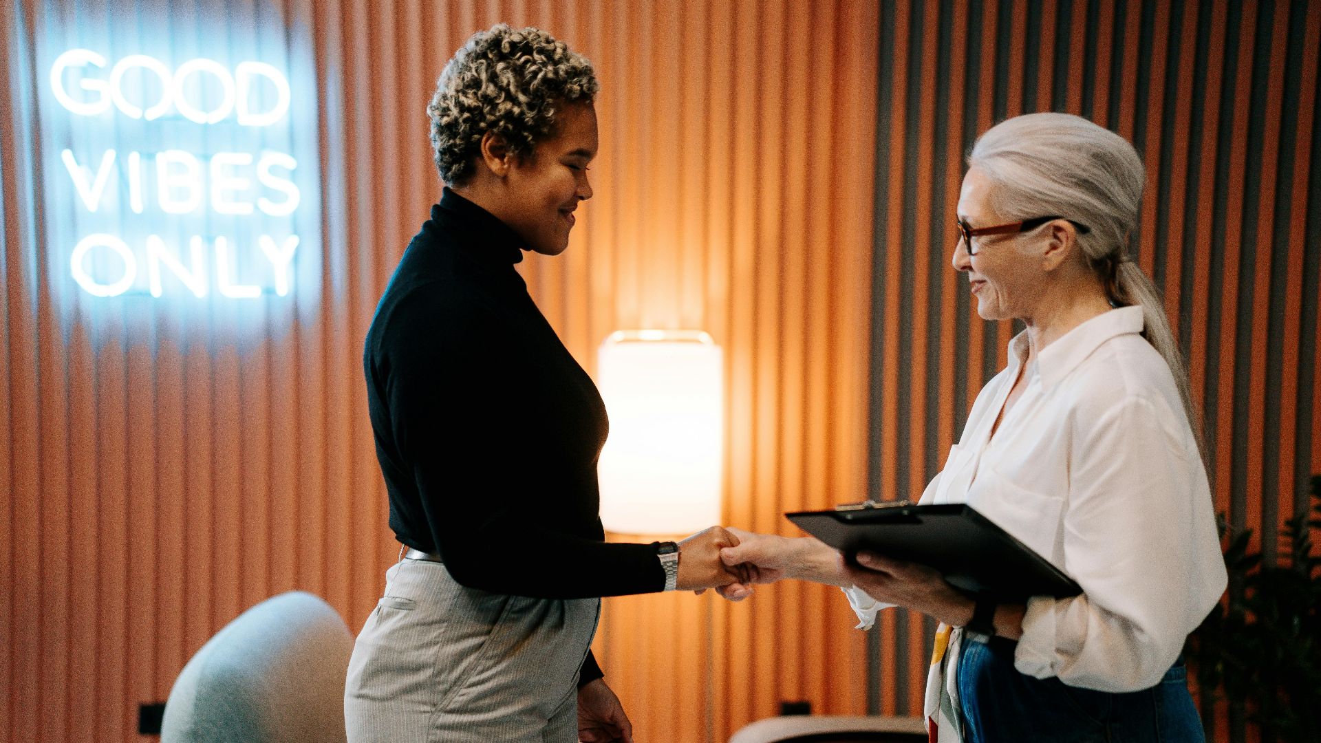 Two diverse businesswomen in a stylish office shaking hands under a 'Good Vibes Only' sign.