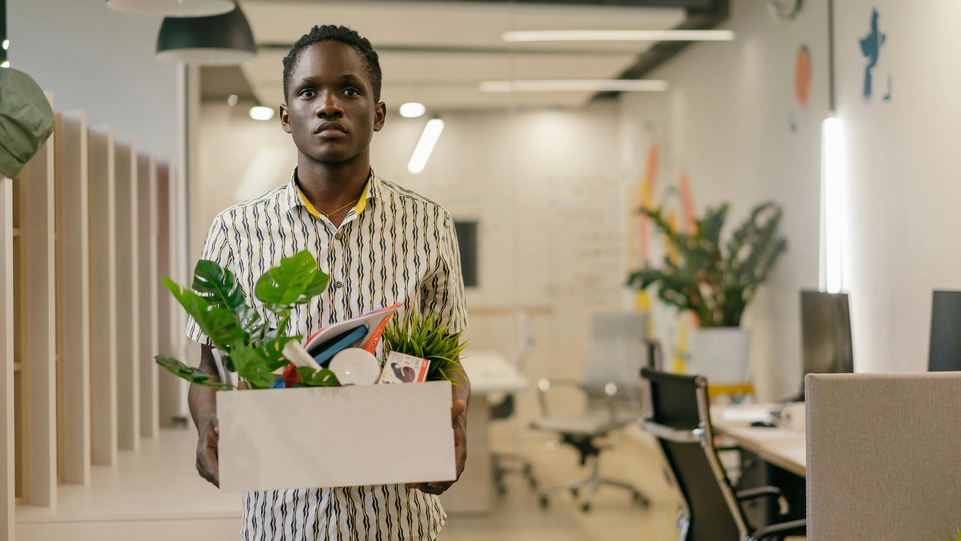 A serious man carries a box of belongings in an office setting, symbolizing dismissal or job change.