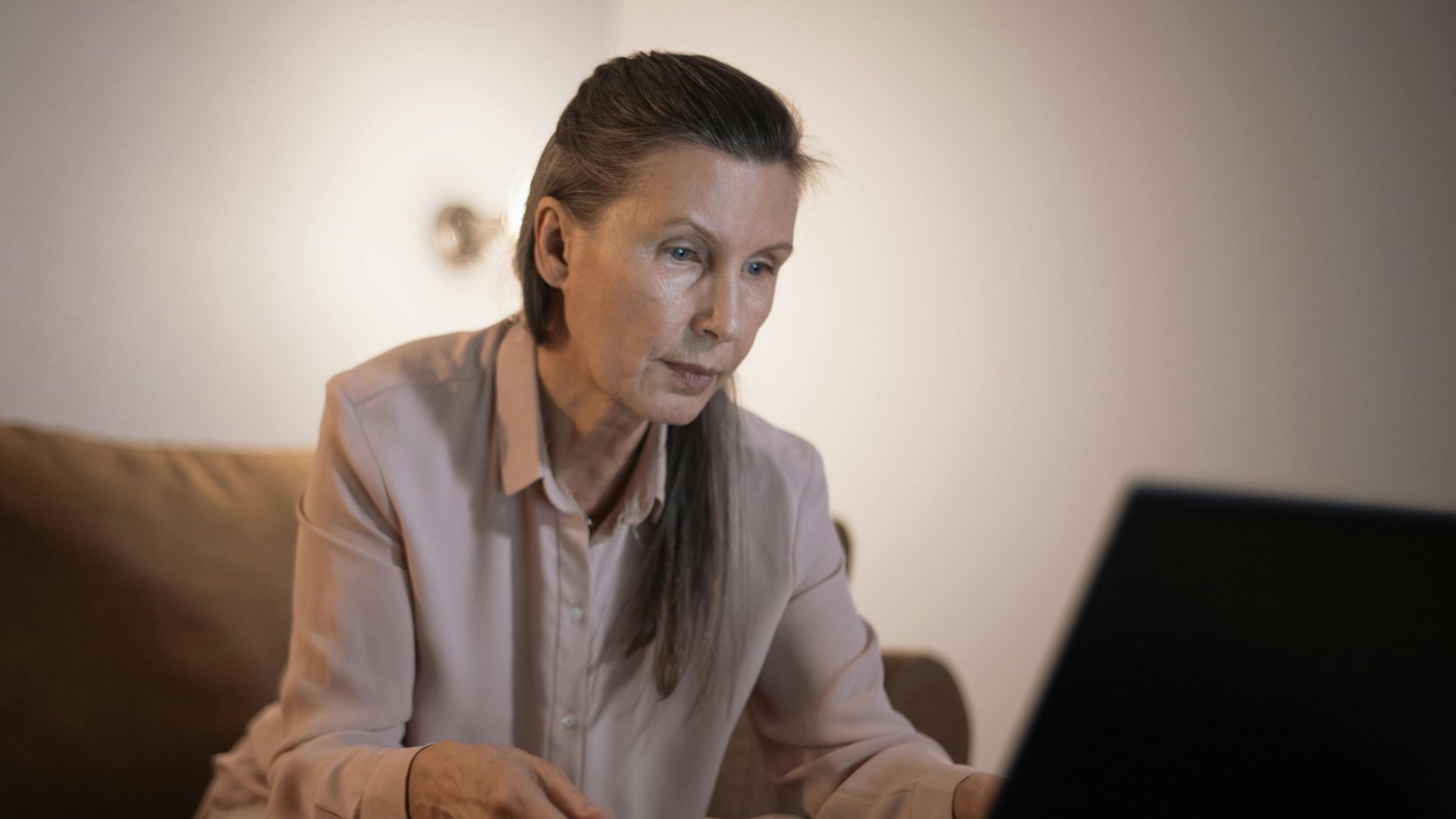 Elderly woman working on laptop in dimly lit living room setting, focused and engaged.