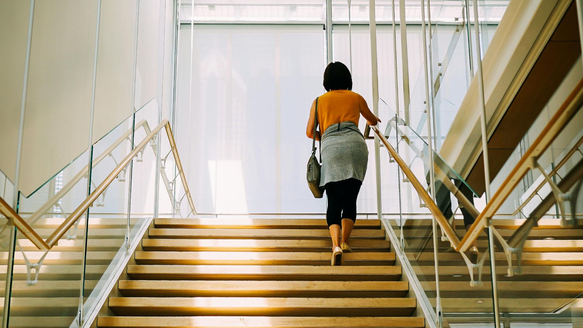A woman walks up a well-lit modern staircase with glass railings inside a building.