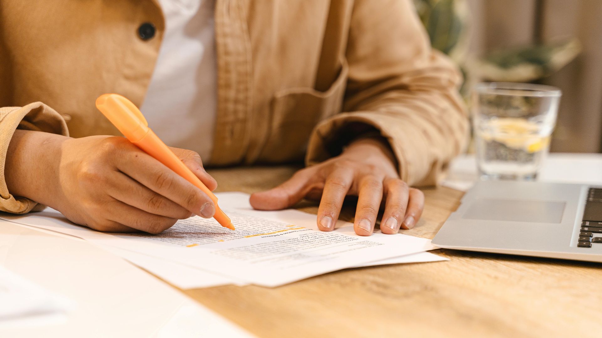 Person highlighting notes at a desk with a laptop and glass of water nearby.