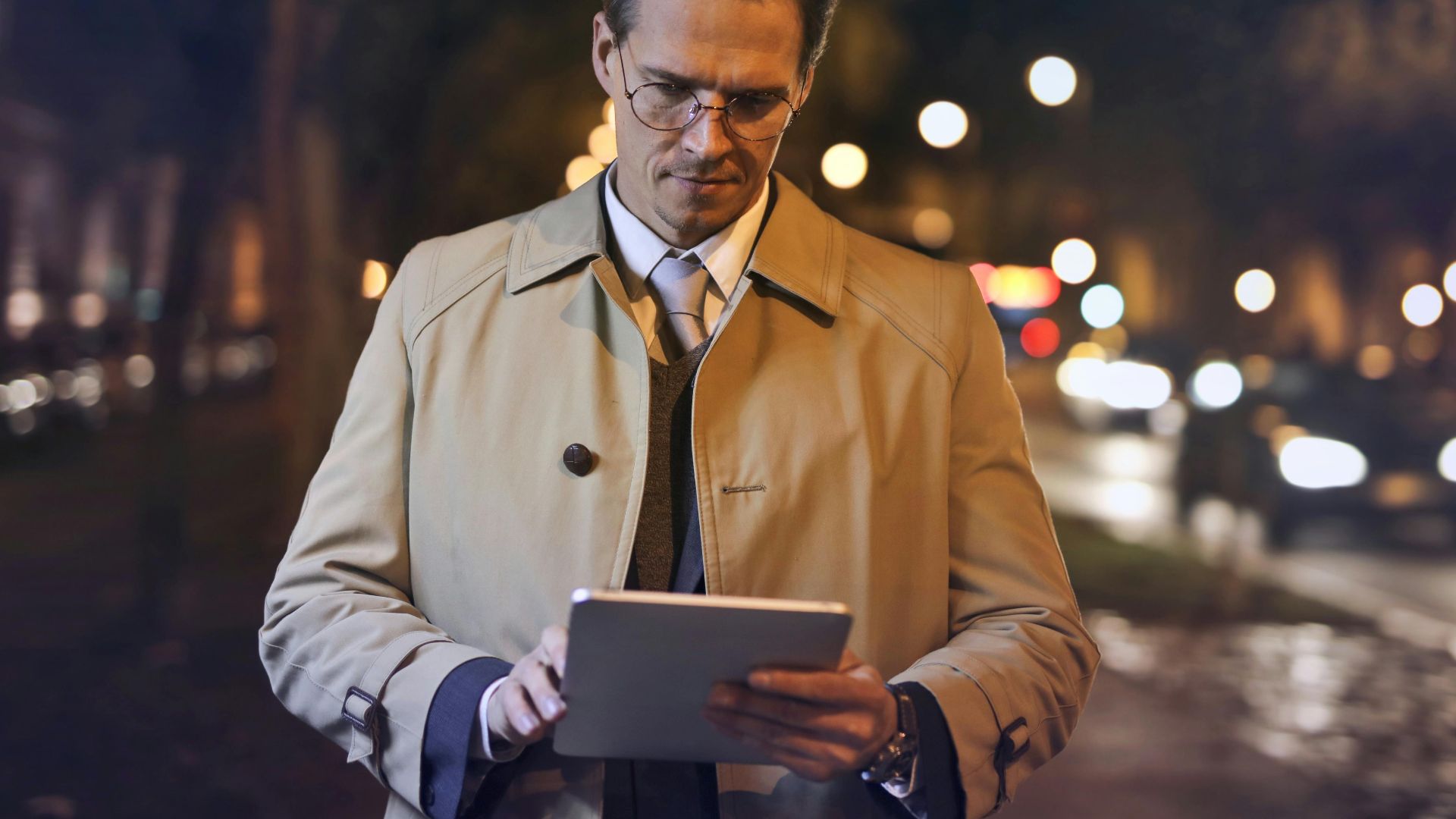A businessman in a trench coat uses a tablet on a city street at night, surrounded by bokeh lights.